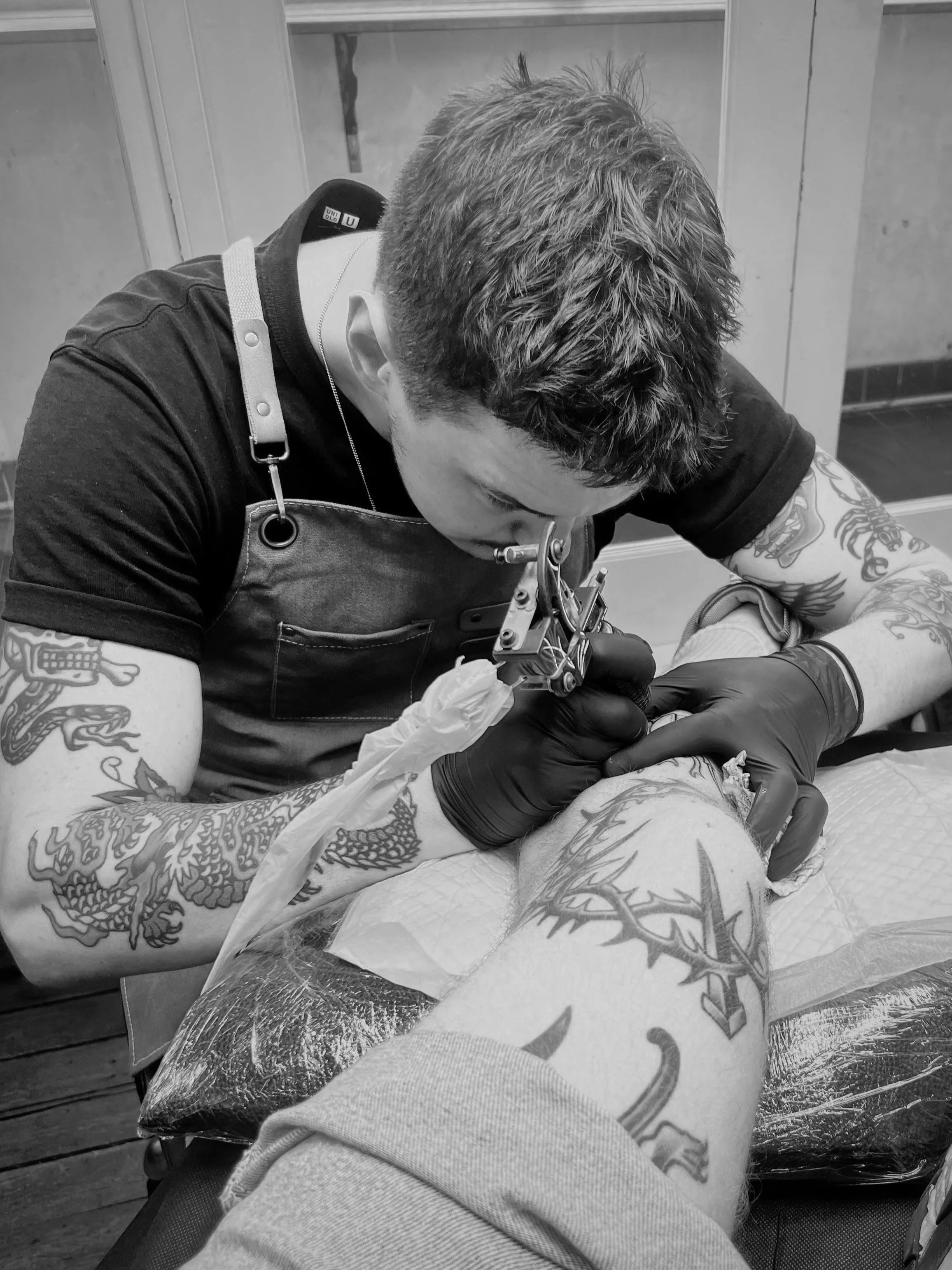 A tattoo artist wearing black gloves, a black shirt, and a leather apron is tattooing a large dragon tattoo on a person's leg in a tattoo studio.