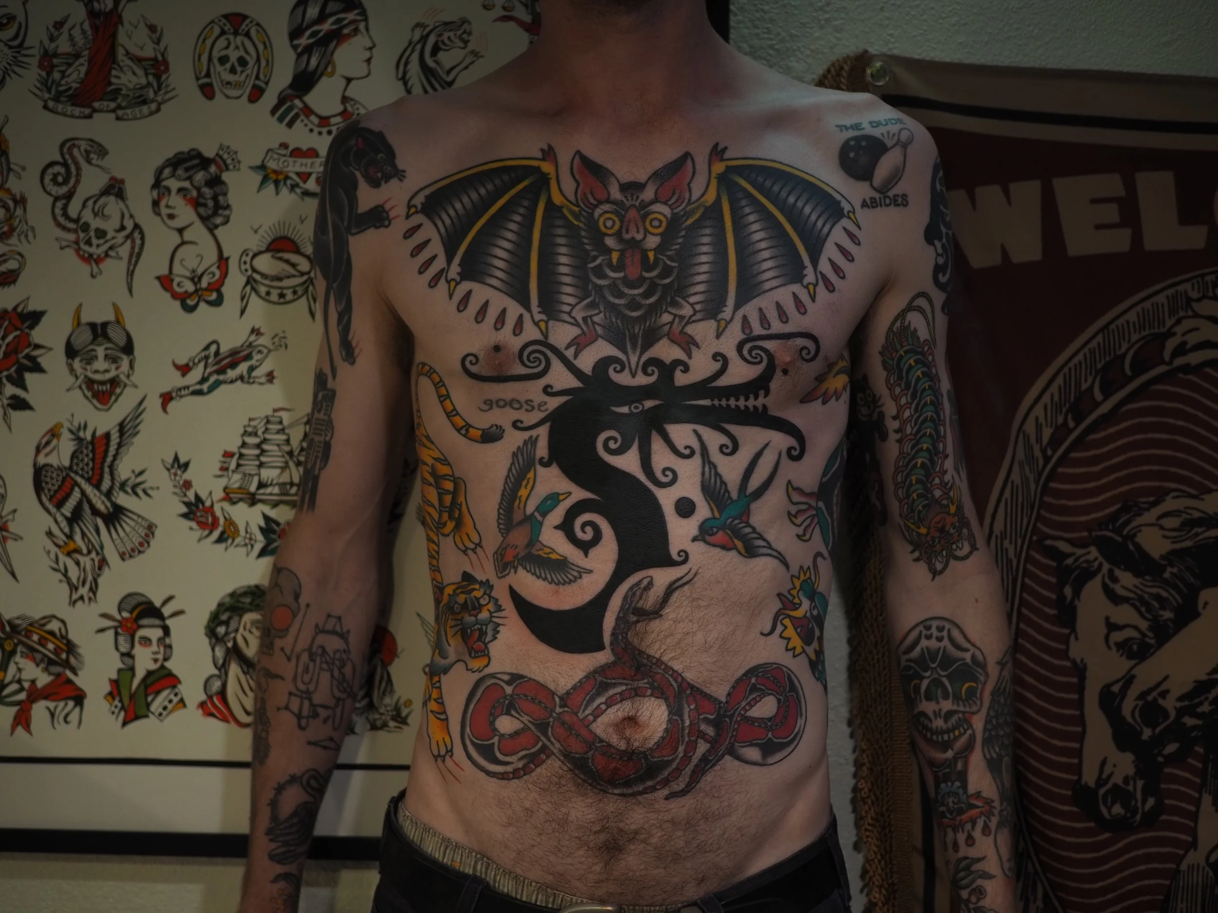 A person's torso with multiple colorful tattoos including a large bat with yellow eyes and outstretched wings, a black and yellow snake, hummingbirds, skulls, and various other designs. The background features tattoo art on the wall.