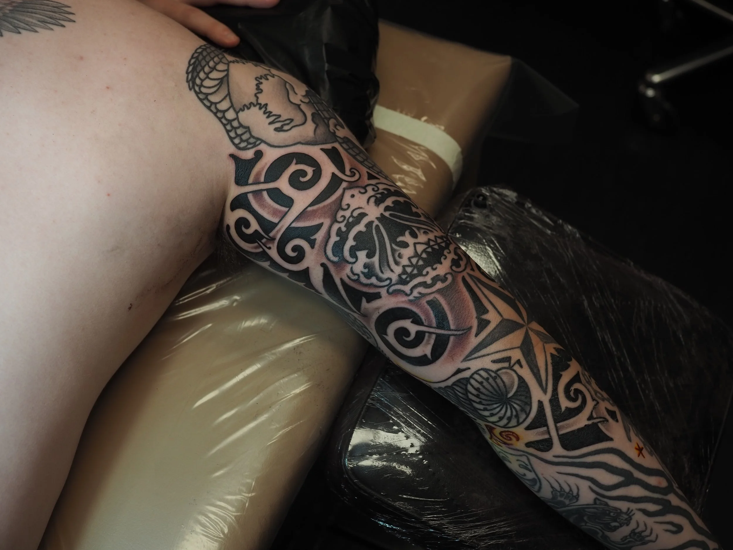 Tattoo artist creating a black tribal-style tattoo on a person's arm, which is resting on a padded surface.