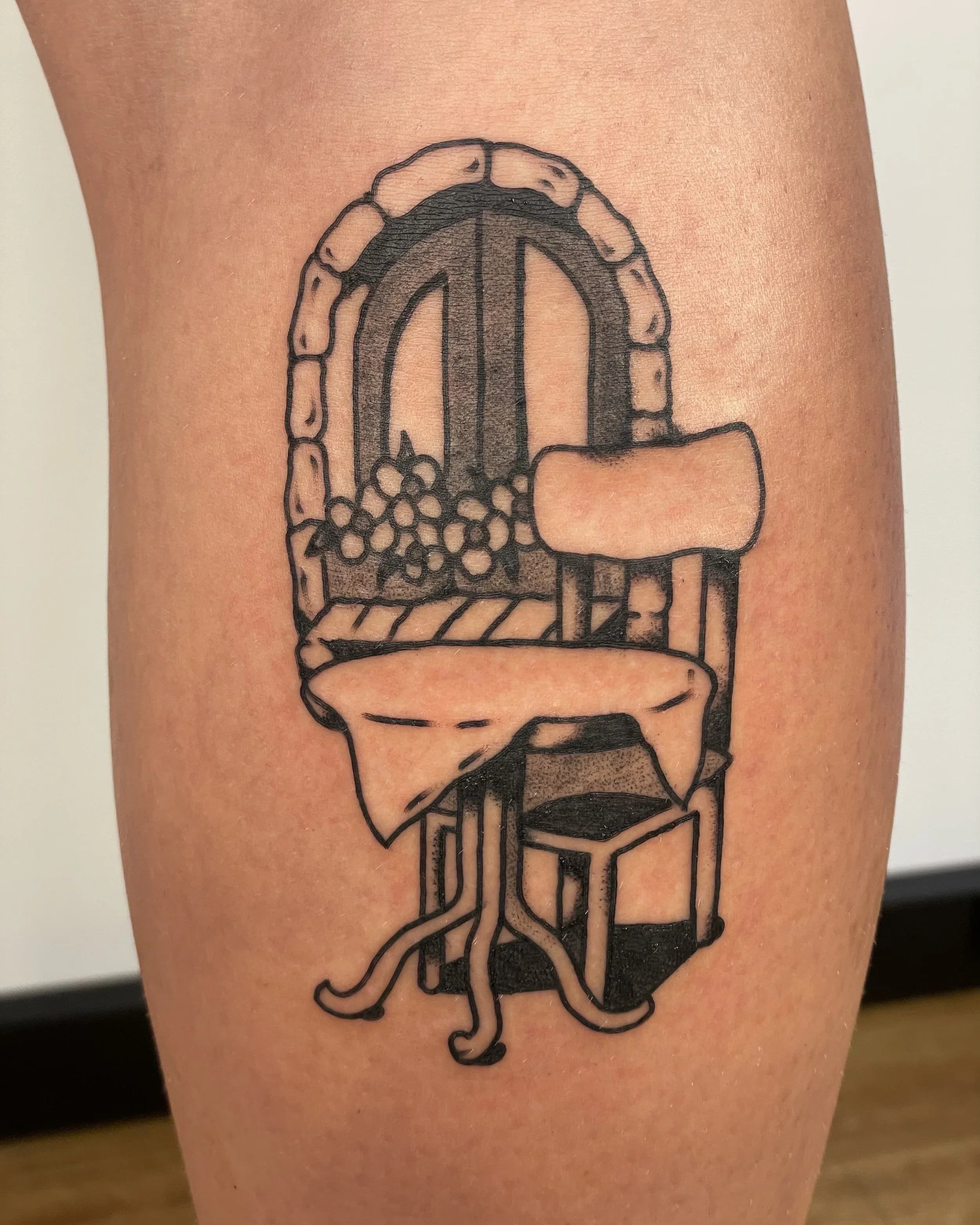 Tattoo of a stained glass window with grapes, a table draped with cloth, and a chair, outlined in black ink.