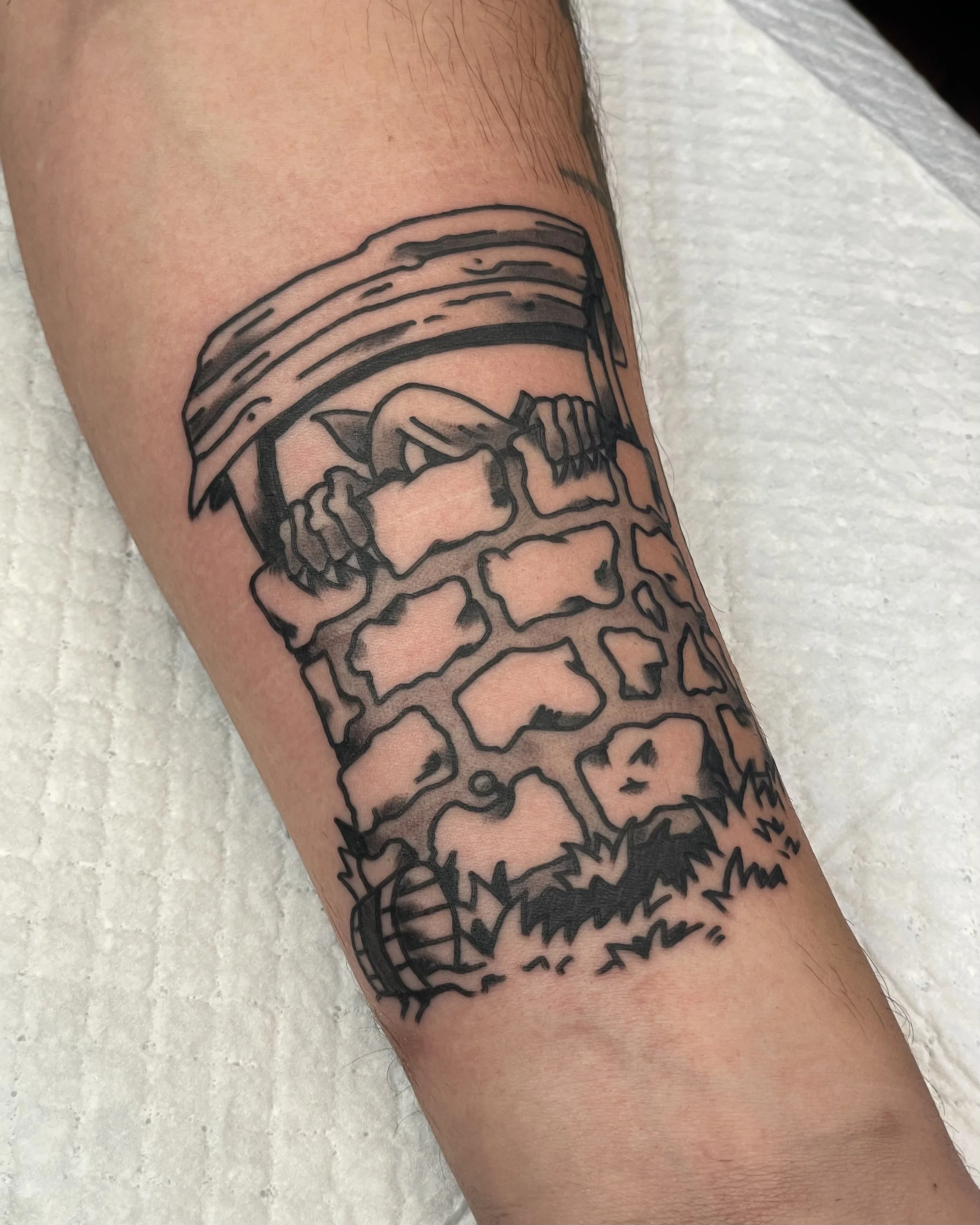Black and grey tattoo of a wishing well and a little goblin hiding inside. Tattood on a forearm.