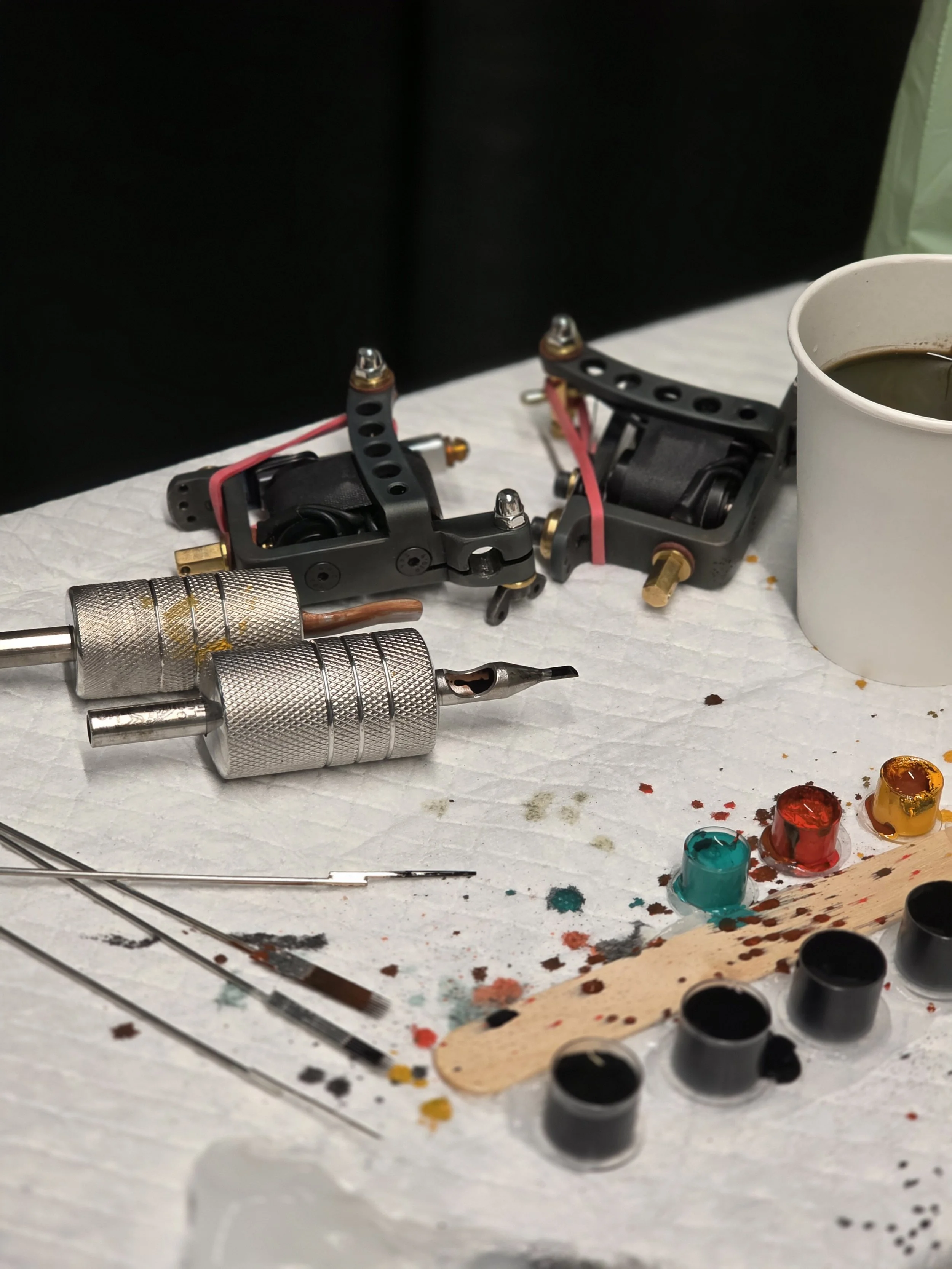 Tattoo equipment including tattoo machines, ink caps, needles, and a cup of coffee on a white paper-covered table.