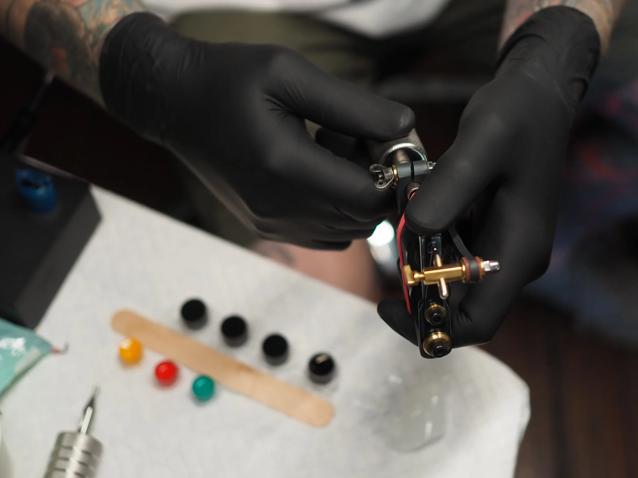 Person wearing black gloves holding a tattoo machine in a studio. Various small parts and tools are on the table, including colored caps and a small file.