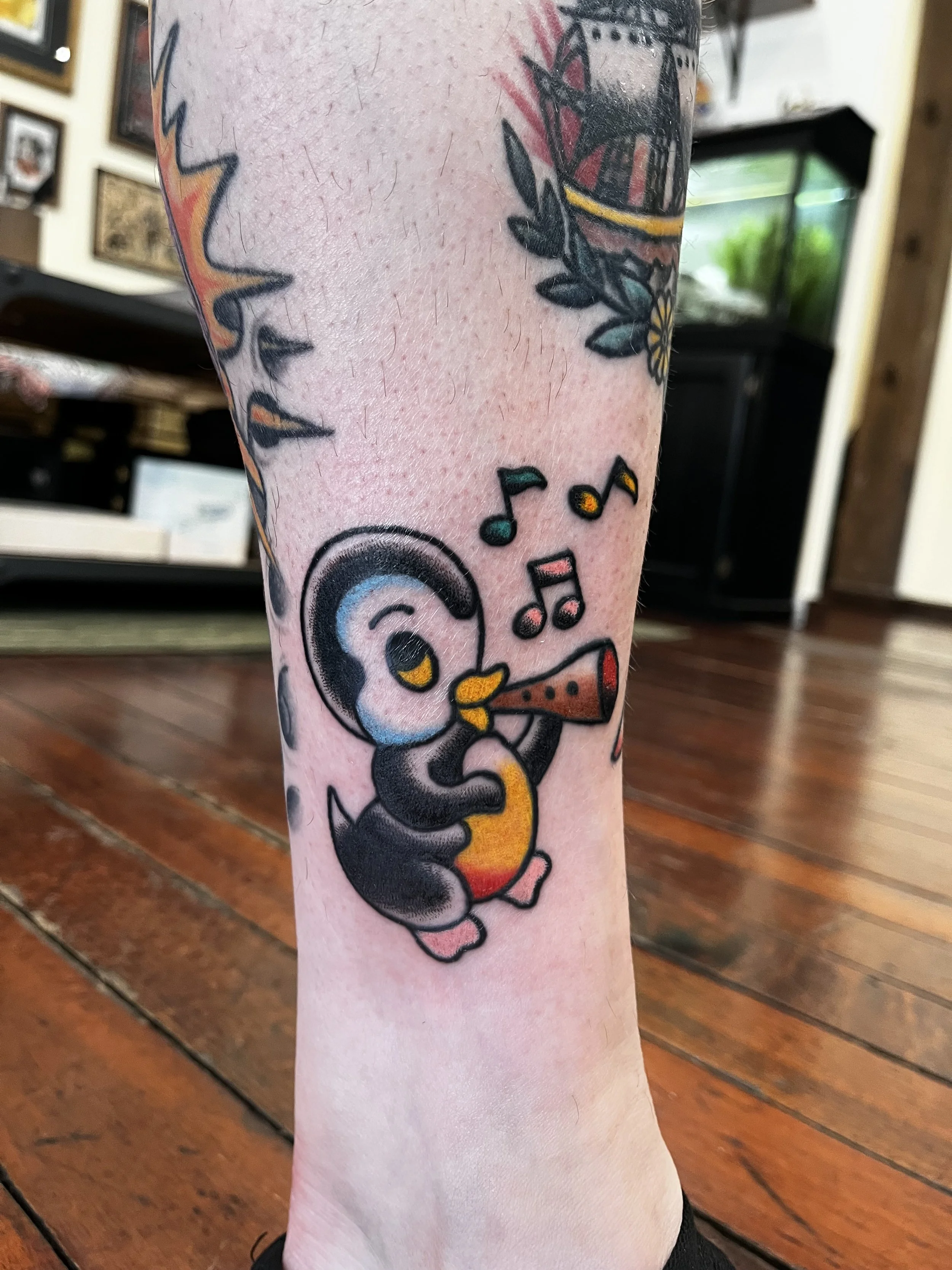 Colorful tattoo of a penguin playing a trumpet, with musical notes around it, on a person's lower leg.