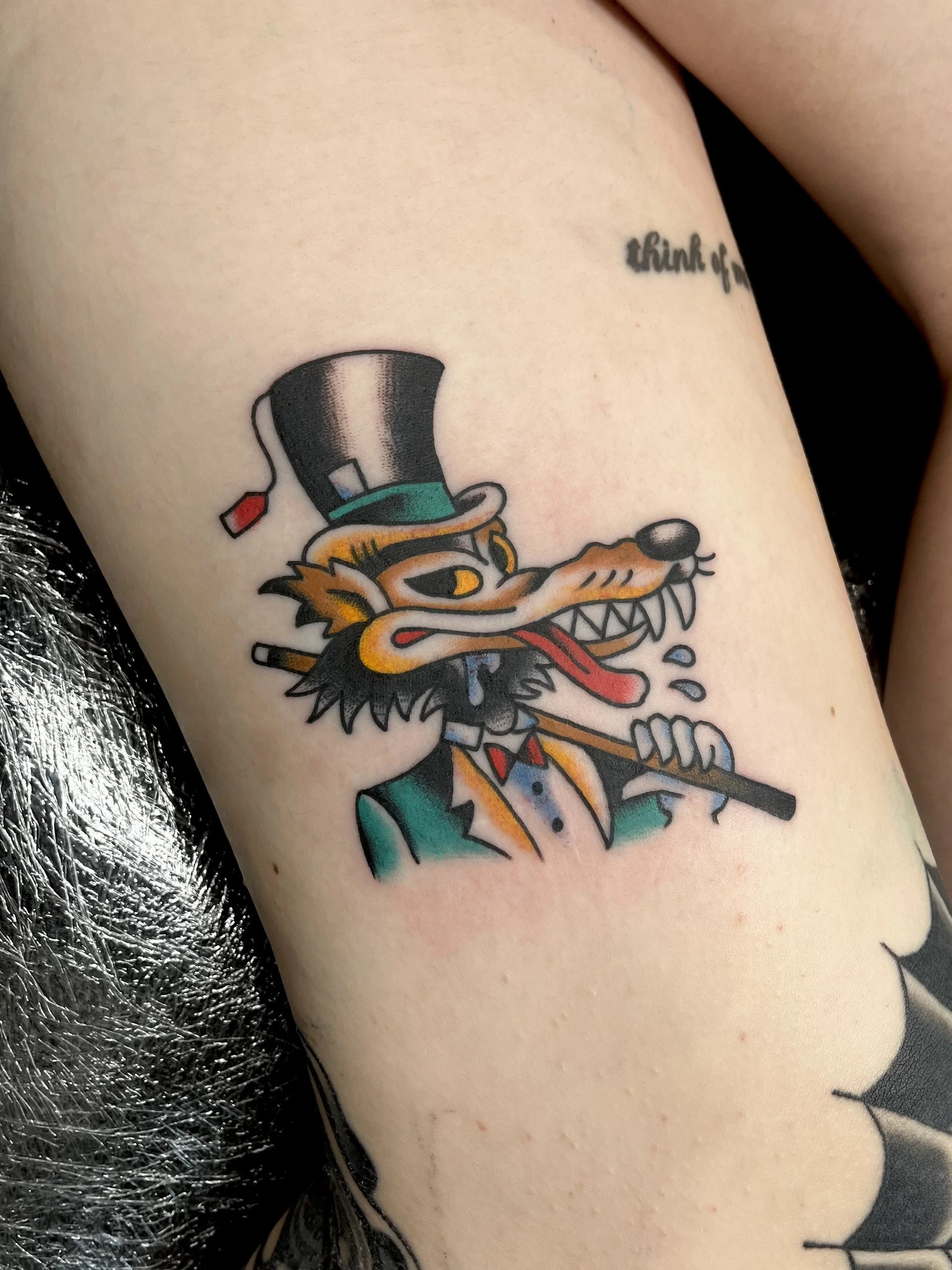 Tattoo of a cartoon wolf dressed in a suit, holding a cane, wearing a top hat with a red feather, and drooling.