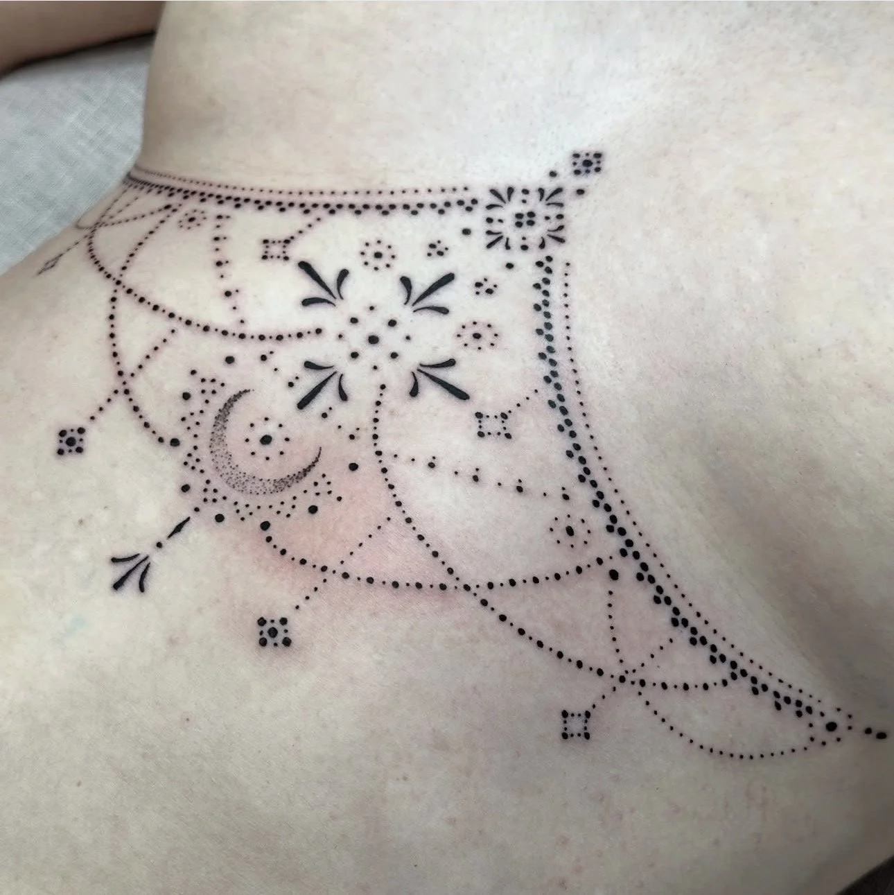 Tattoo on person's collarbone area featuring a dotted, geometric, floral, and celestial pattern with a crescent moon and decorative elements.