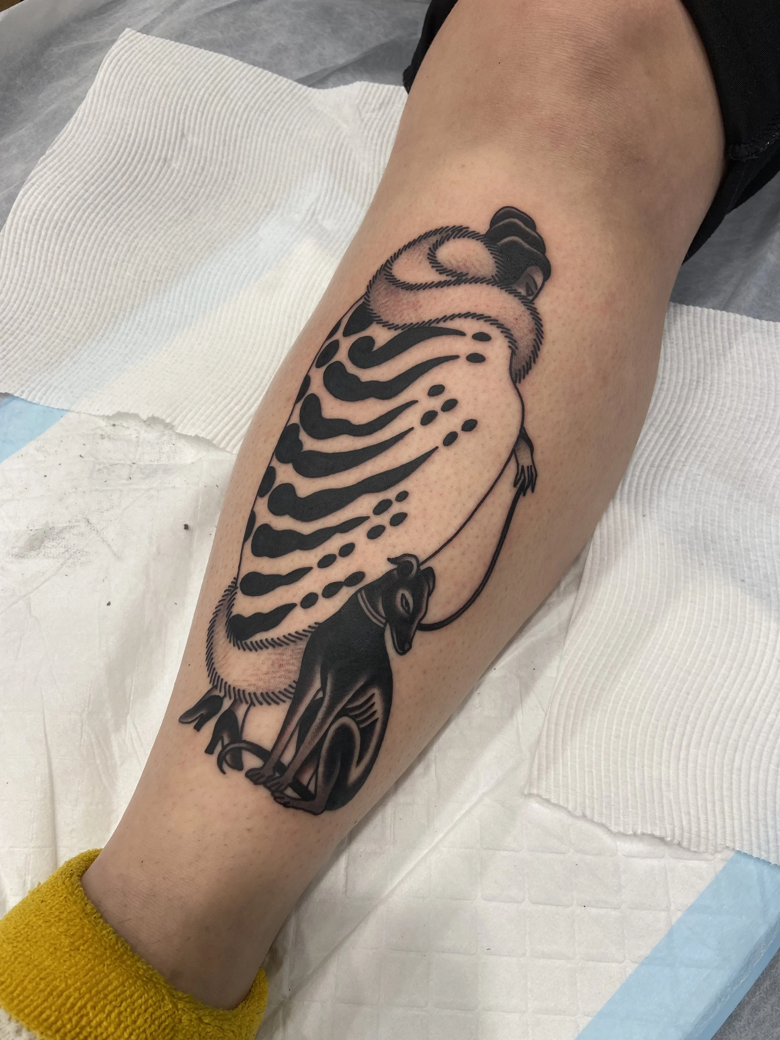 Black and gray tattoo of a woman wrapped in a cloth with a tie around her waist, holding a leash attached to a portrait of a dog, on a person's thigh.