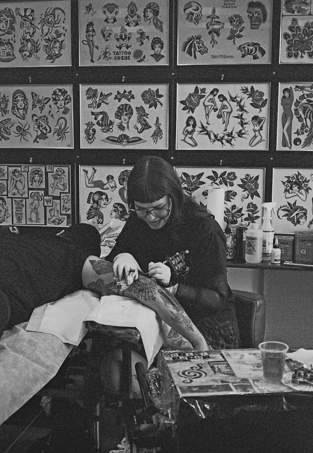 A woman doing a tattoo on a person's arm in a tattoo shop, with tattoo flash art on the wall behind her.