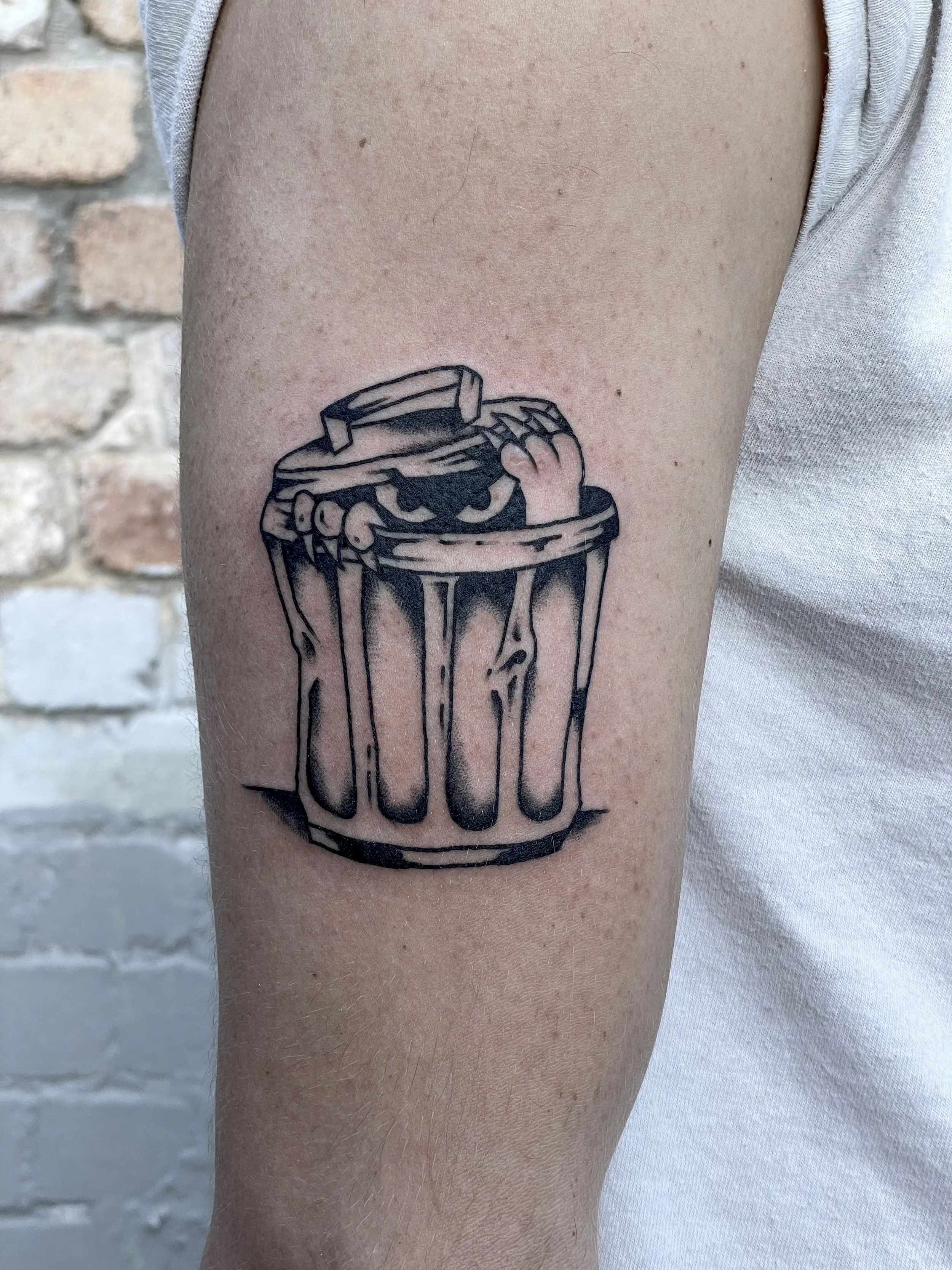 Tattoo of a trash can with a pair of hands reaching out from inside, depicted in black ink with a cartoonish style.