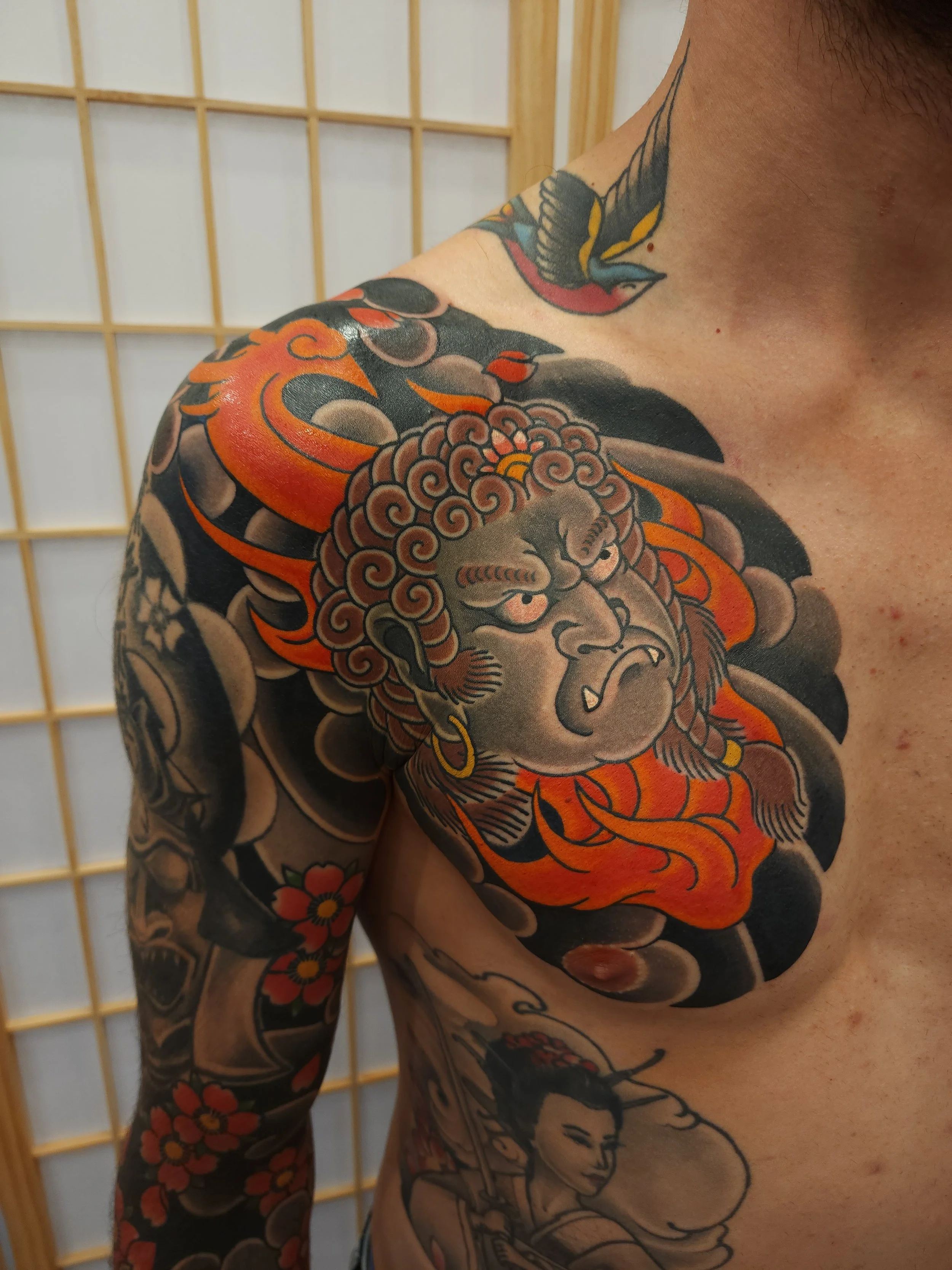 Tattoo of a fierce fudo myoo with flames, and clouds on a person's shoulder, with additional floral and figure tattoos on the arm and chest.