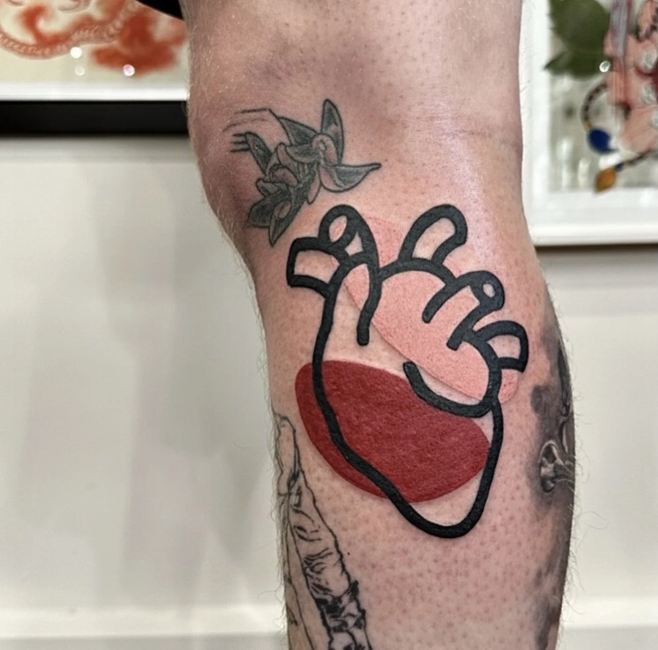 Tattoo of a stylized human heart with a red fill and black outline on a person's arm, with additional tattoos visible nearby.