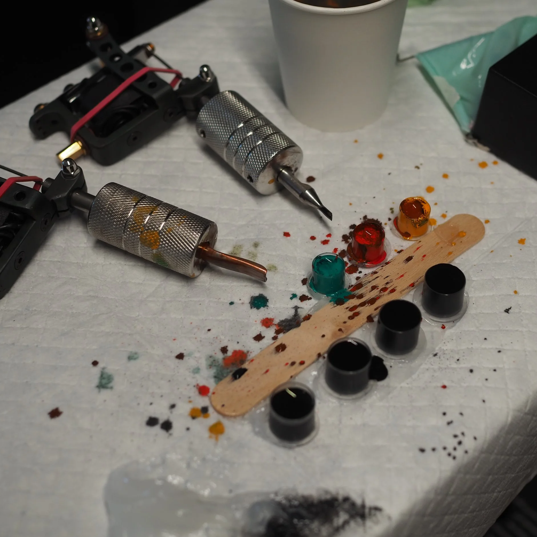 A collection of tattooing supplies including tattoo machines, ink caps filled with colored inks, ink pots, and a wooden palette with spilled ink, all arranged on a paper-covered surface.
