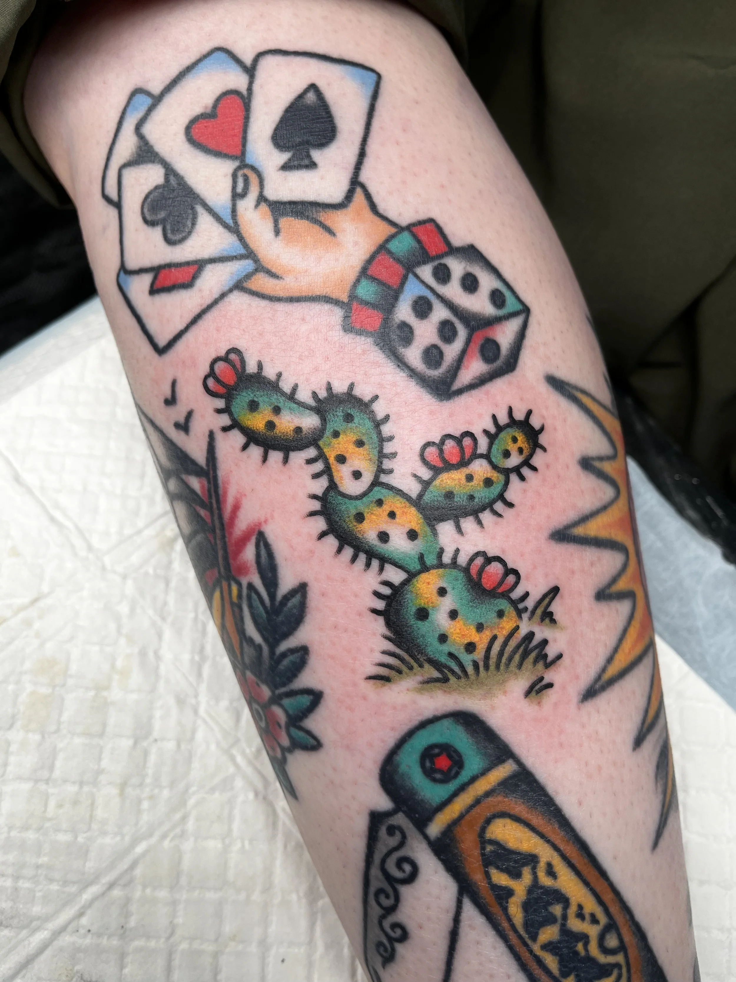 Tattoo featuring a hand holding four playing cards, a colorful cactus plant, a rolling pin, and a zigzag line. The tattoo is in a traditional style with bold outlines and vibrant colors.
