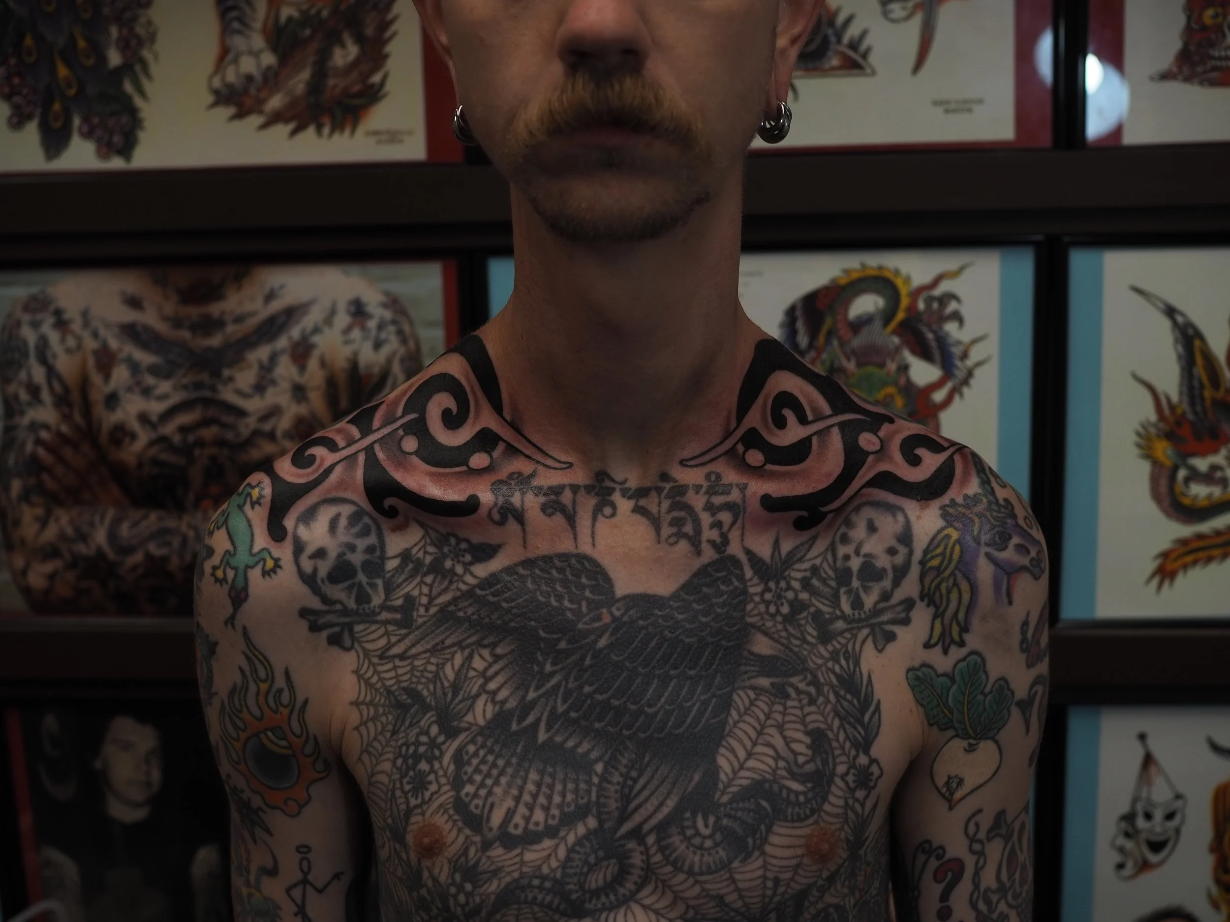A shirtless man with tattoos on his chest and arms, with a large black eagle tattoo in the center, skull and crossbones, and other colorful designs. He has earrings and a mustache, standing in front of tattoo artwork on the wall.