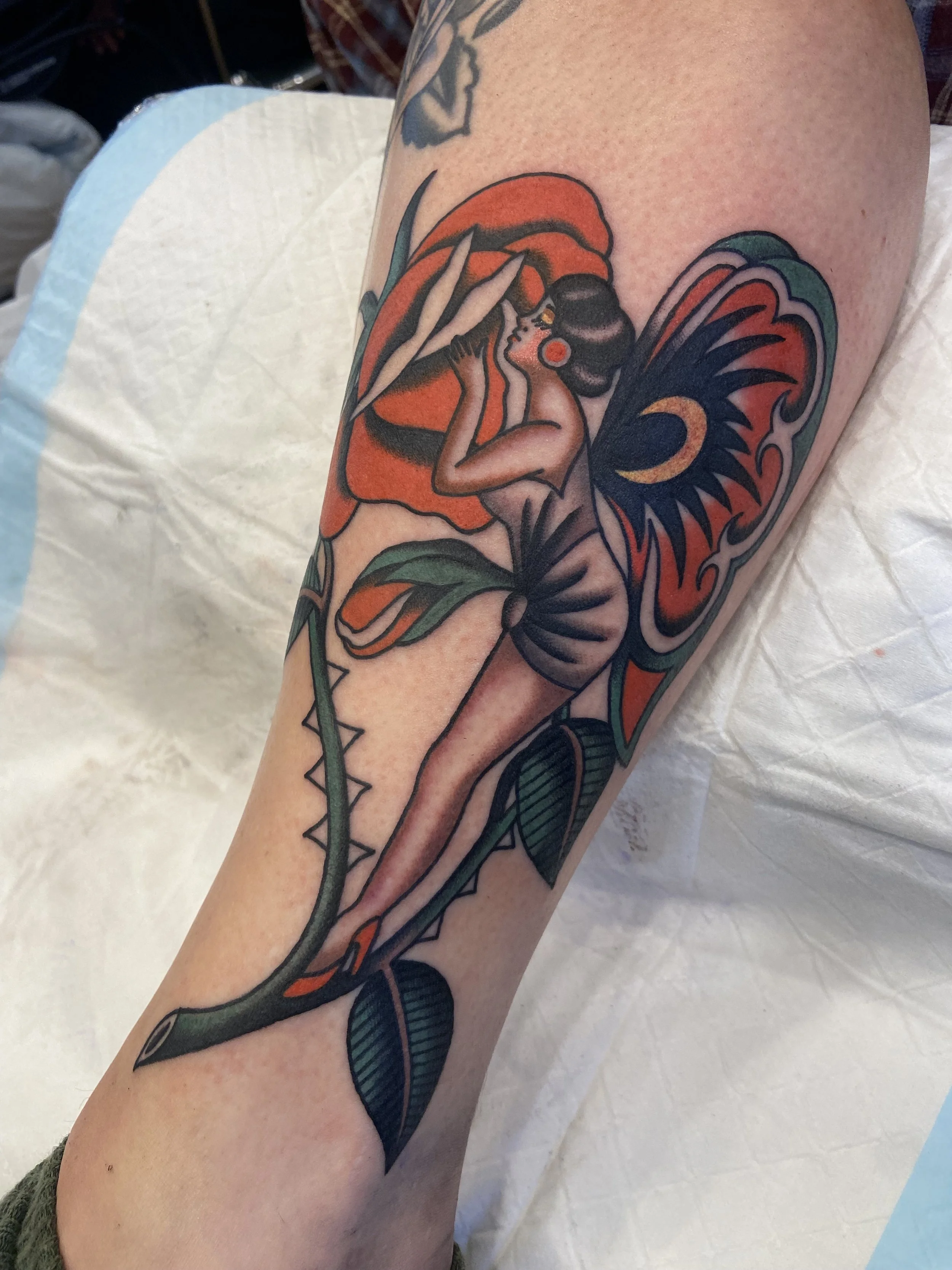 Tattoo of a stylized woman with butterfly wings sitting on a rose and a thorny stem, with green leaves and bold colors.