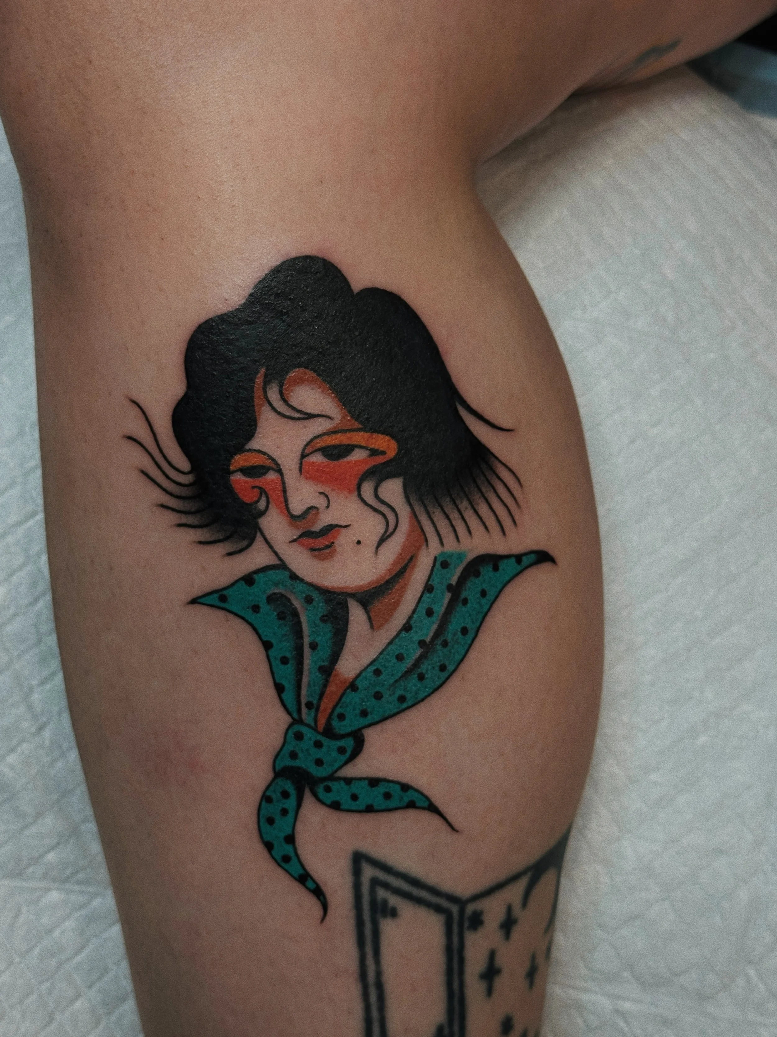 Tattoo of a woman with short black hair, orange eye makeup, and a blue polka-dot scarf on the upper arm.