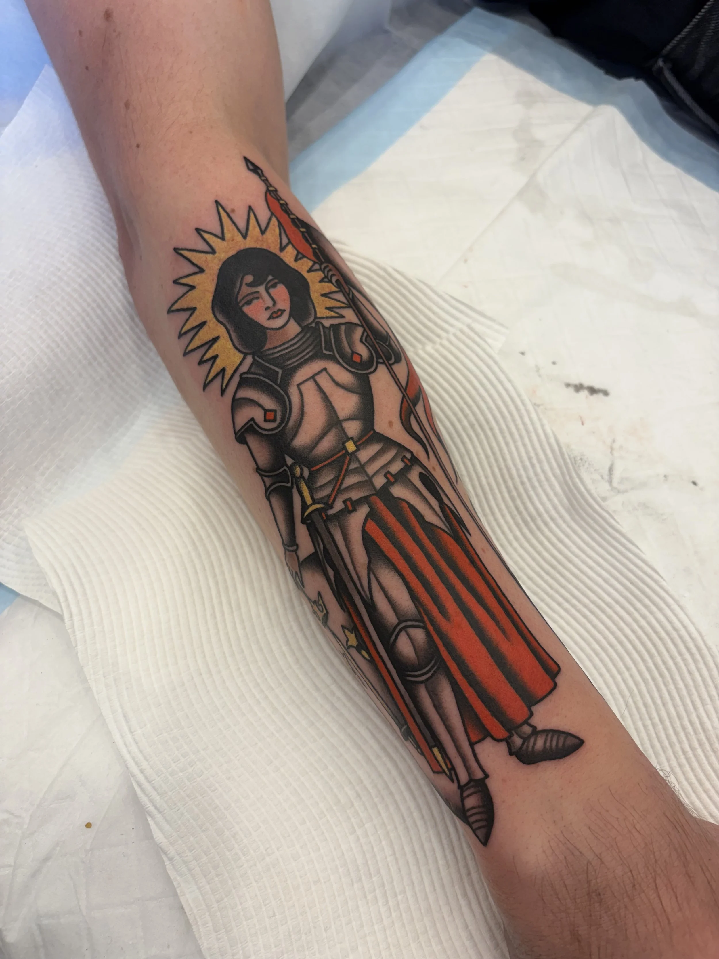 Tattoo of a woman in armor holding a spear, with a yellow sunburst behind her head, and a red cape. She has dark hair and a serious expression.