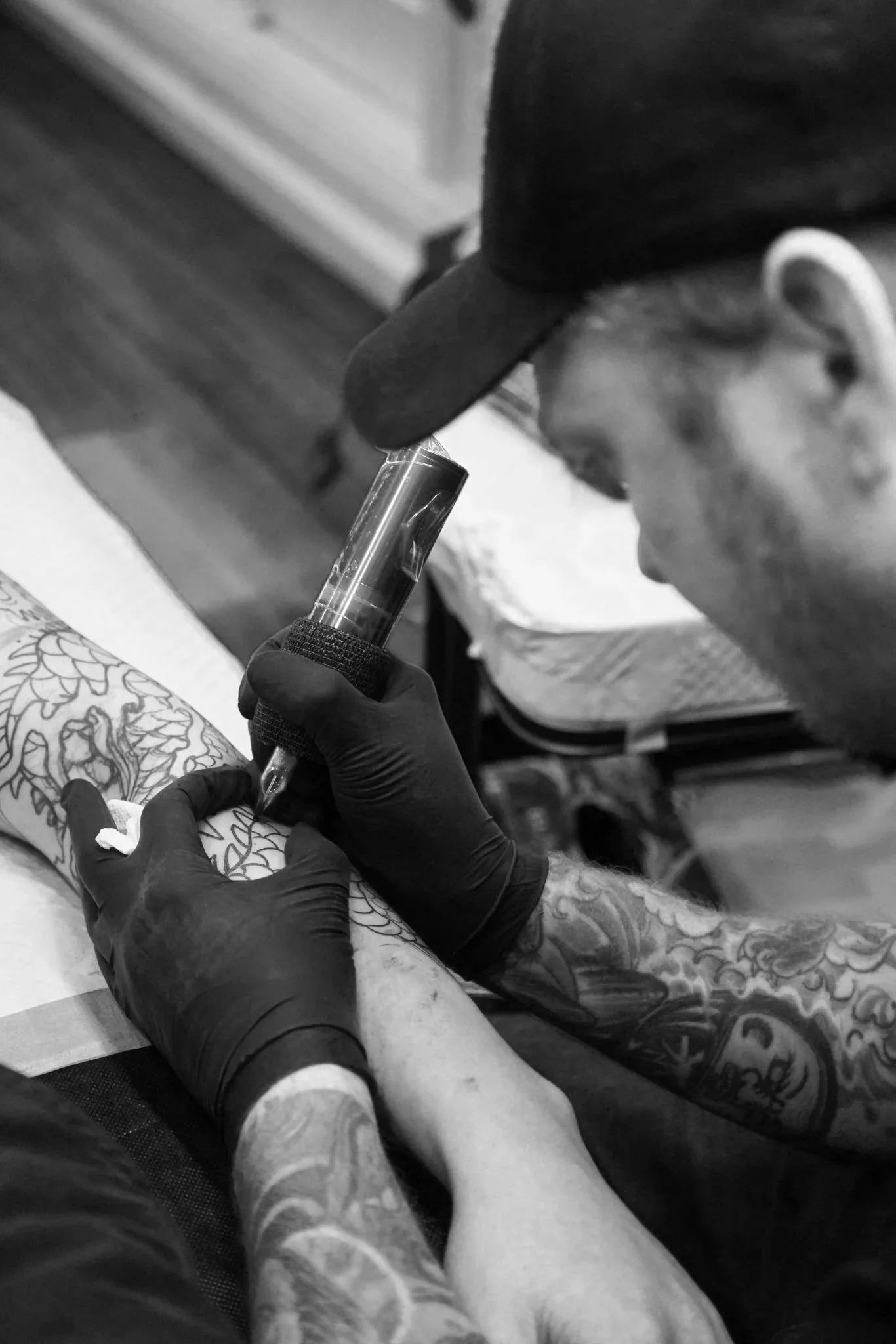 Tattoo artist wearing black gloves and a black cap ink tattoos on a person's arm using a tattoo machine in a tattoo studio.