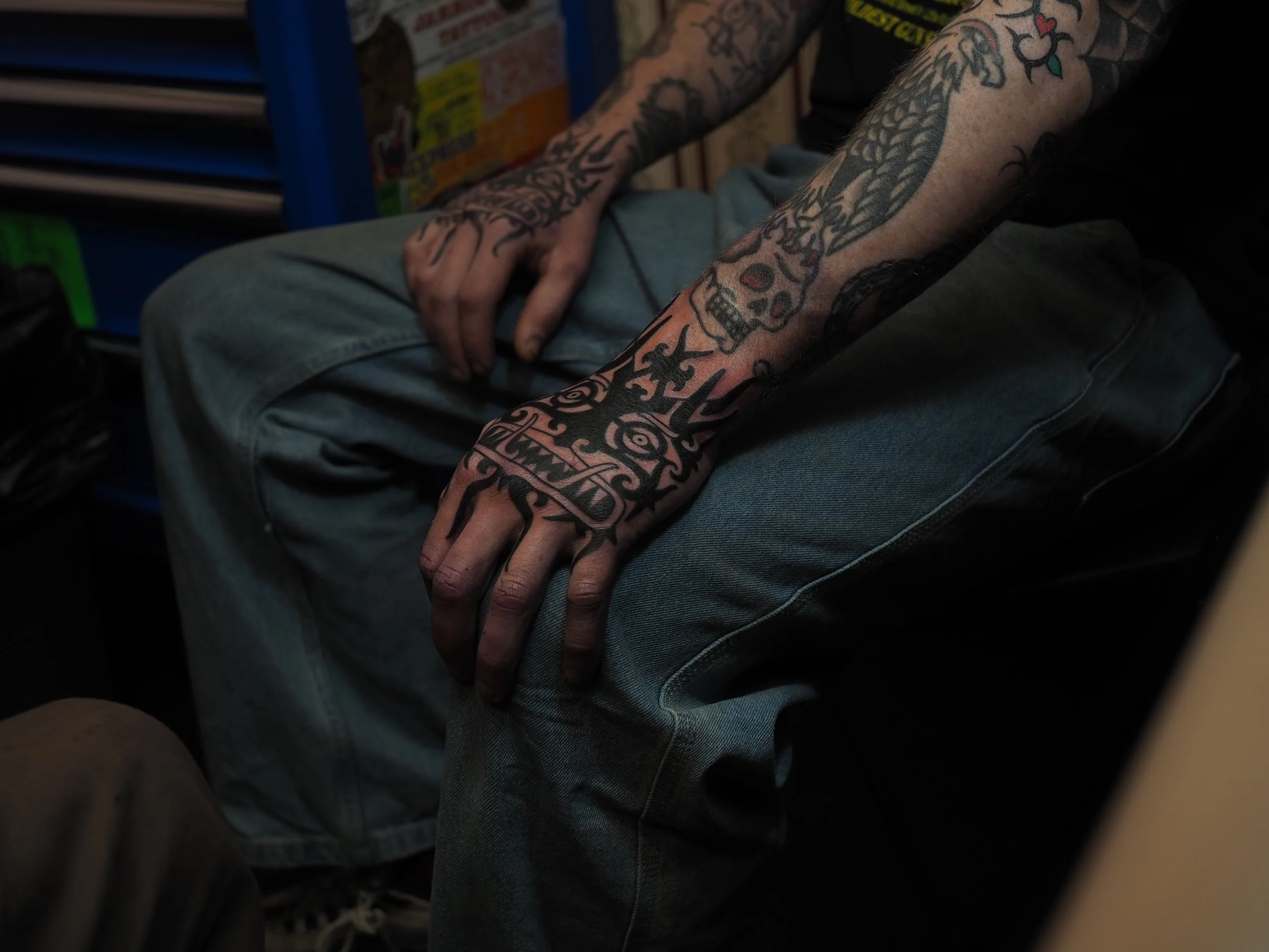 Close-up of a person's arm and hand resting on their knee. The person has multiple tattoos, including a dragon or snake on the forearm, a skull with a helmet, and tribal patterns on the hand.