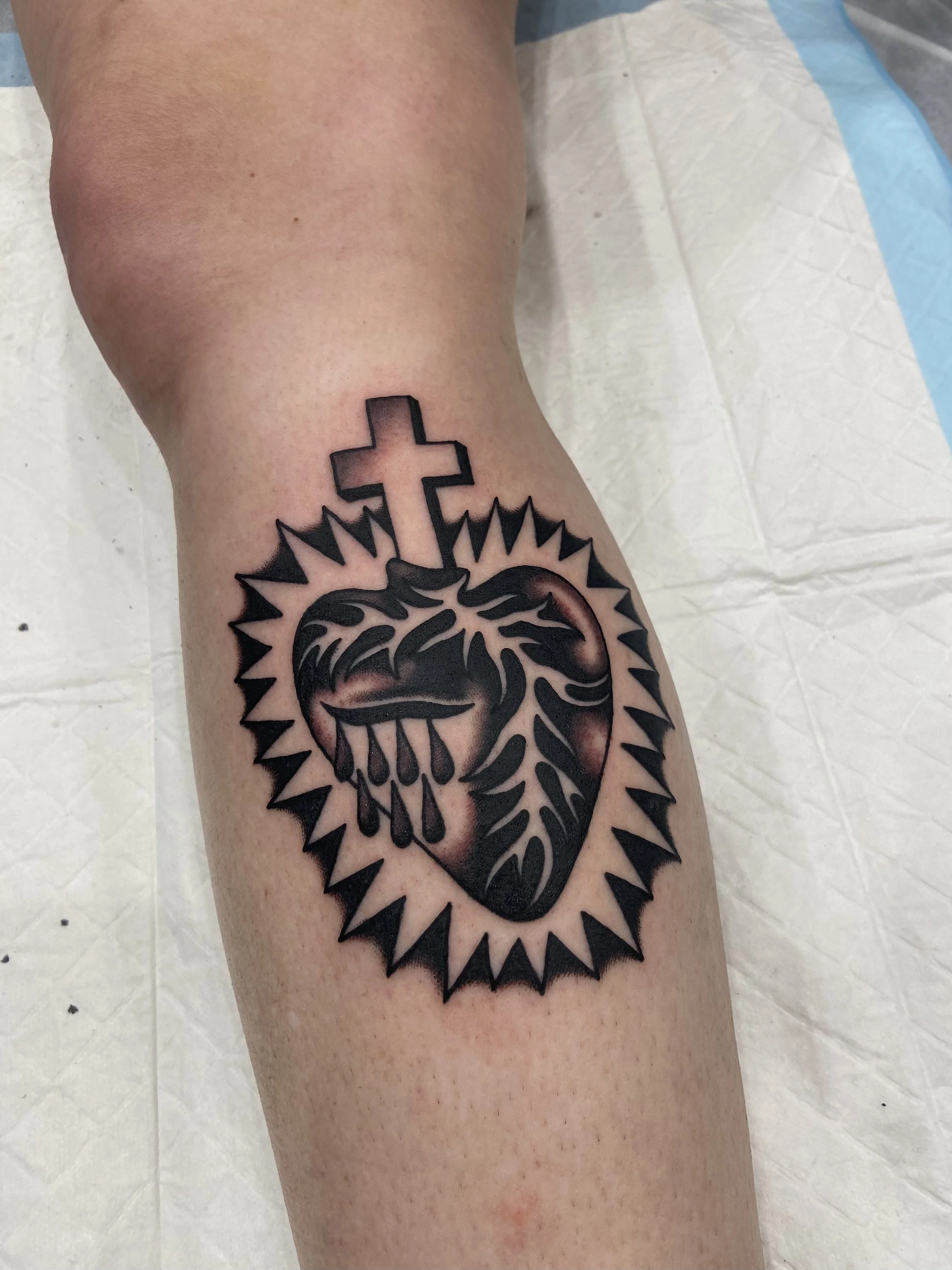 Tattoo of a bleeding heart with a crown of thorns, a cross above, and a radiant outline around it on a person's leg.