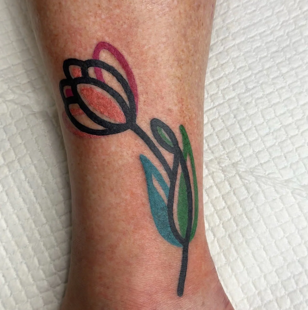 Colorful tattoo of a stylized flower with black outline, pink, green, and blue accents on a person's arm.