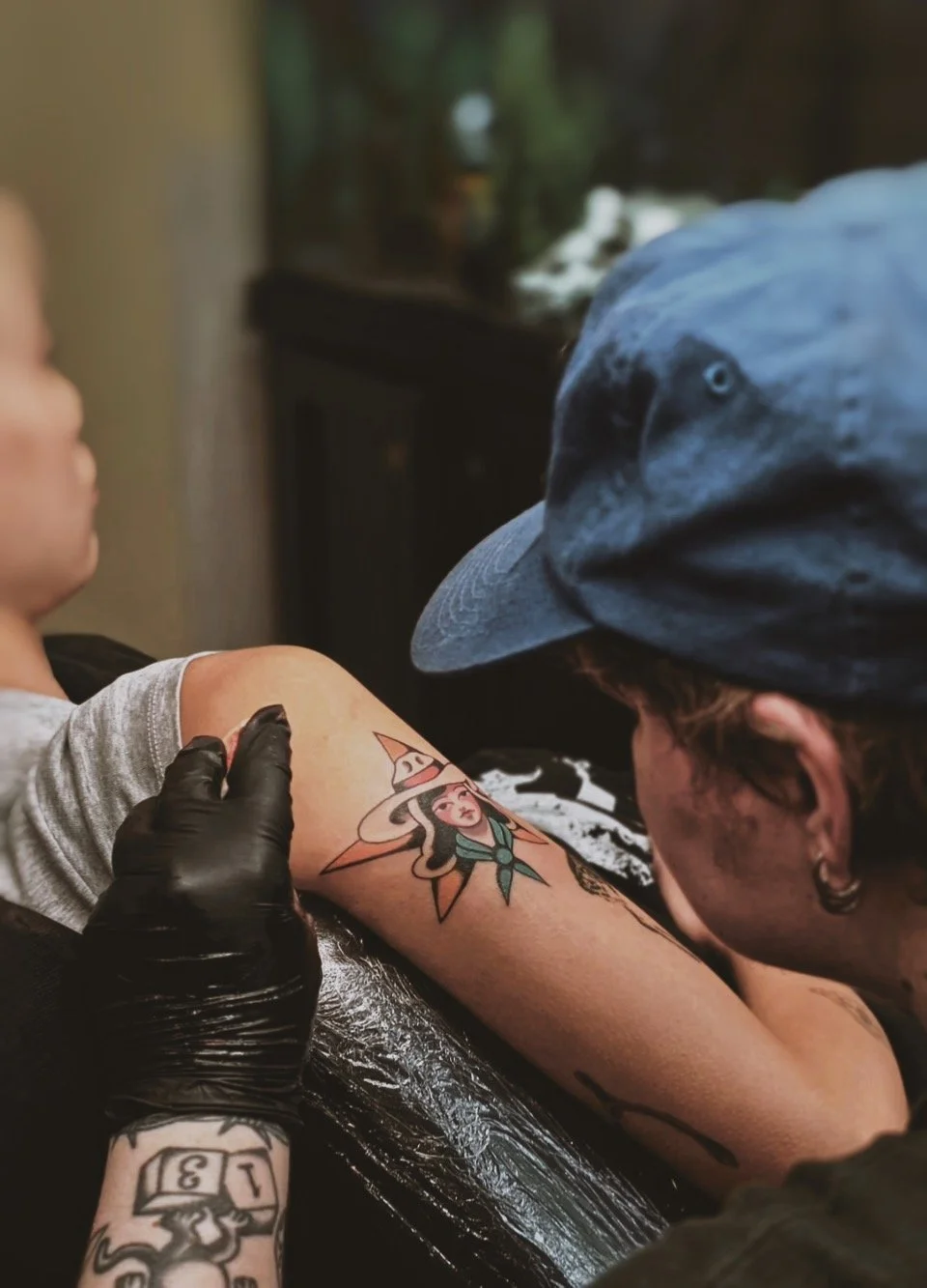 A person getting a tattoo of a witch wearing a pointed hat on their arm at a tattoo parlor, with a tattoo artist working on it.