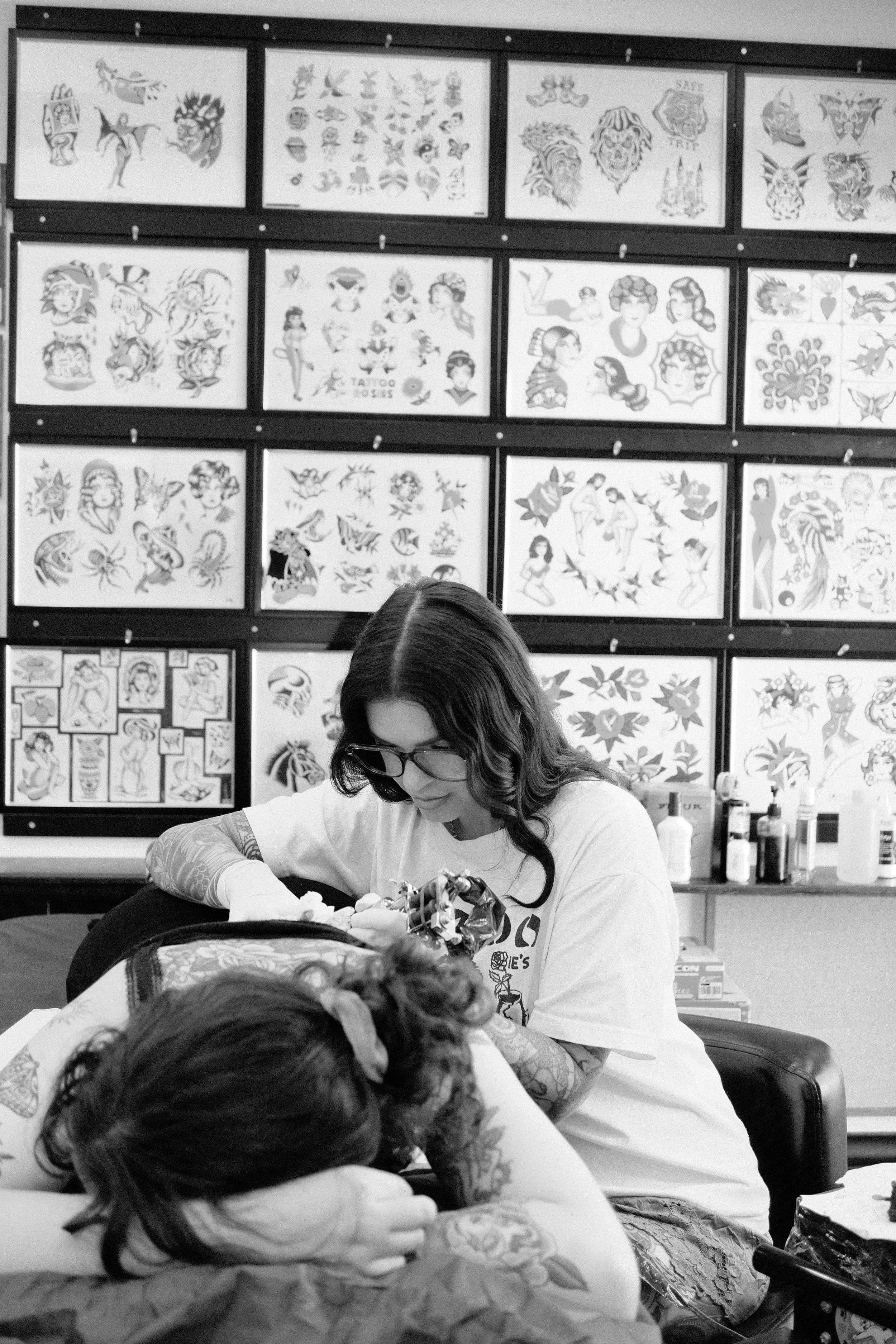 A woman doing a tattoo on a person's arm in a tattoo parlor, with tattoo artwork displayed on the wall behind her.