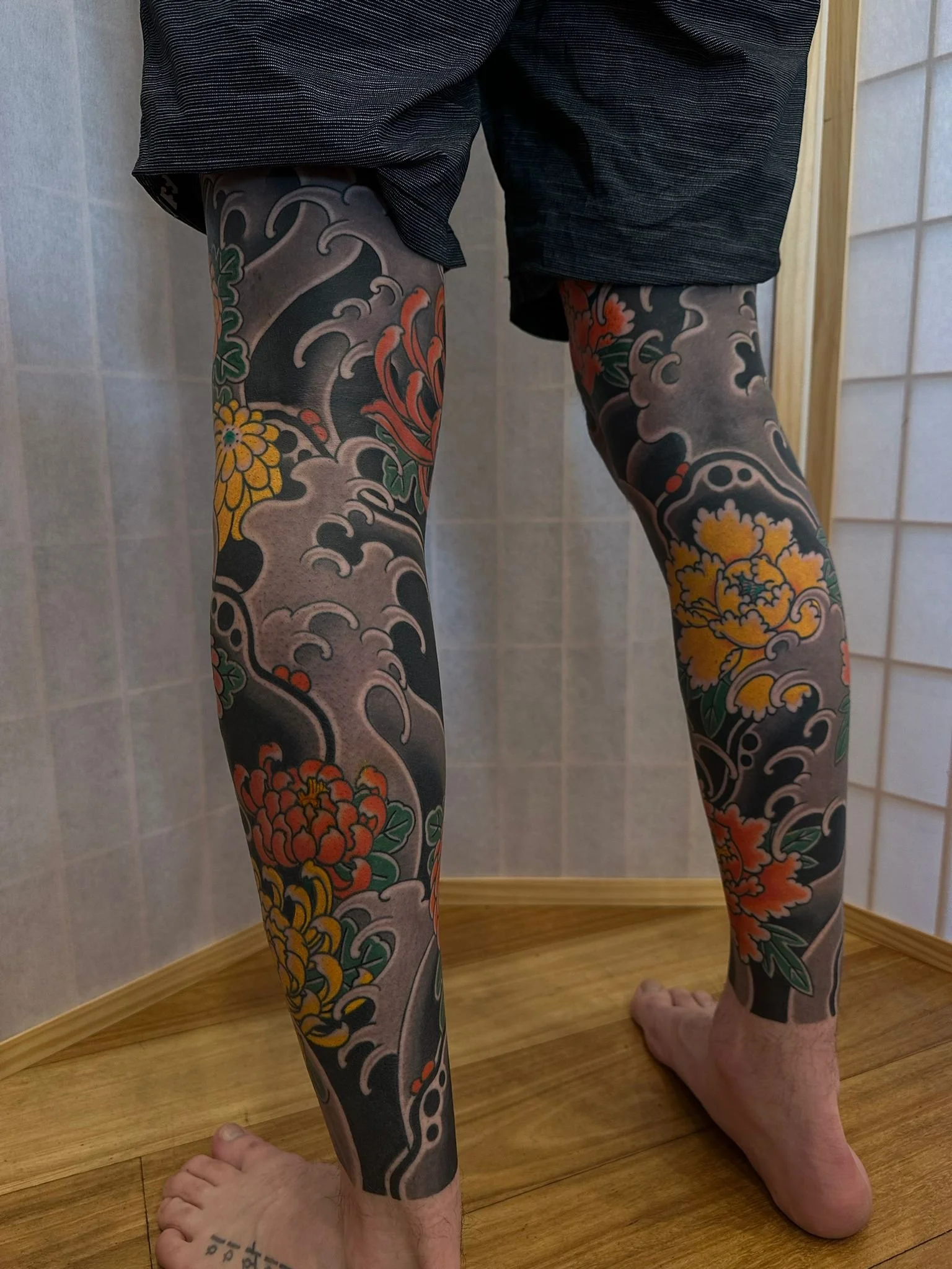Person showing colorful traditional Japanese-style tattoos on both legs, standing in a room with wooden flooring and tiled walls.