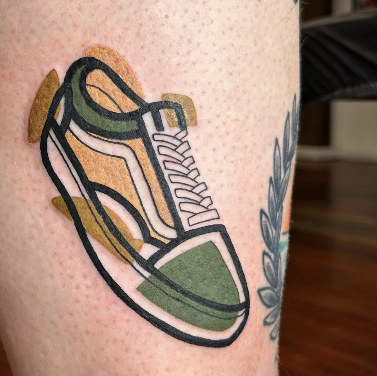 Tattoo of a stylized sneaker on skin with red freckles, next to a leafy branch.