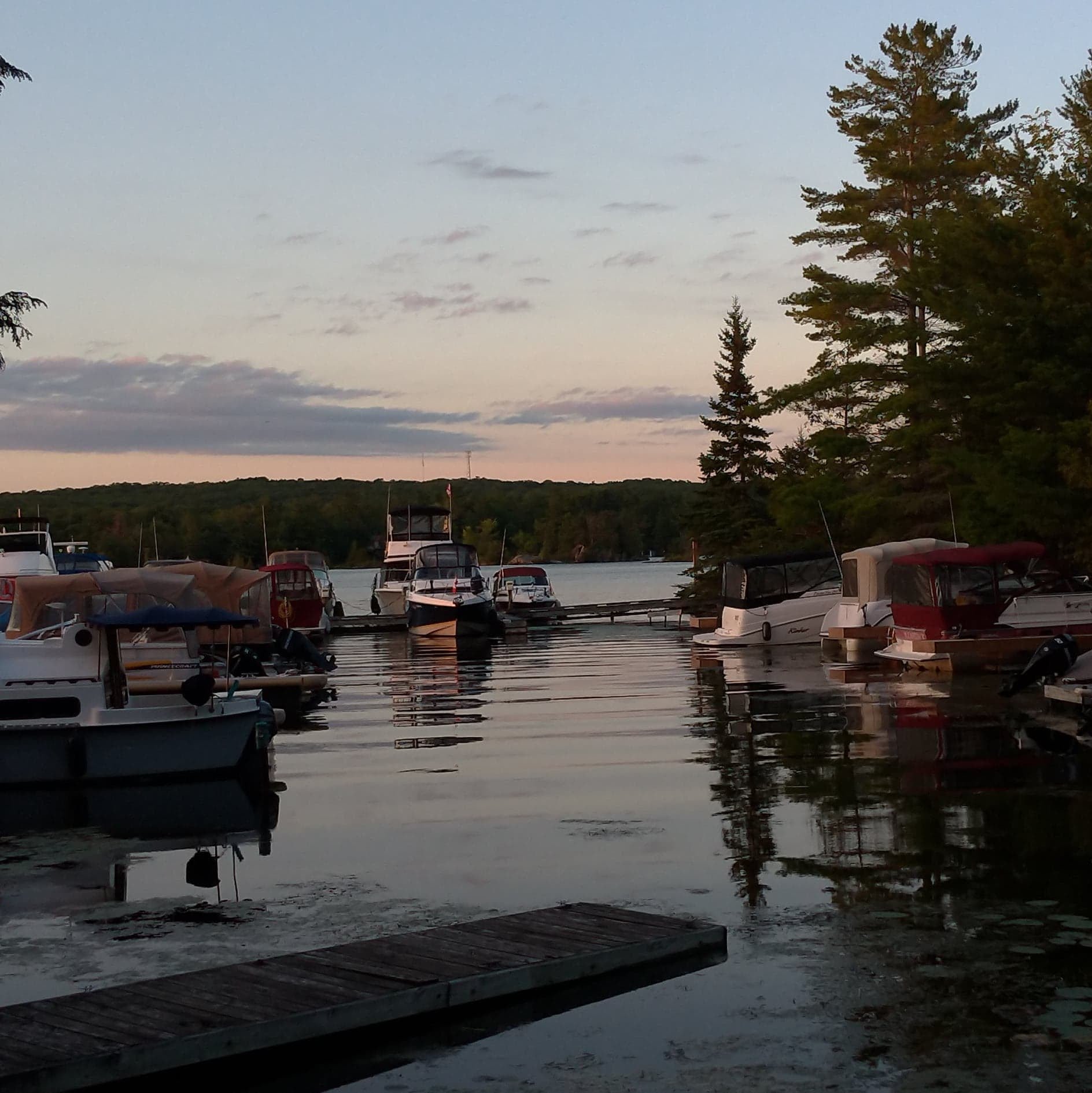 Boats docked at a serene lake dock during evening with calm water, trees, and a cloudy sky.