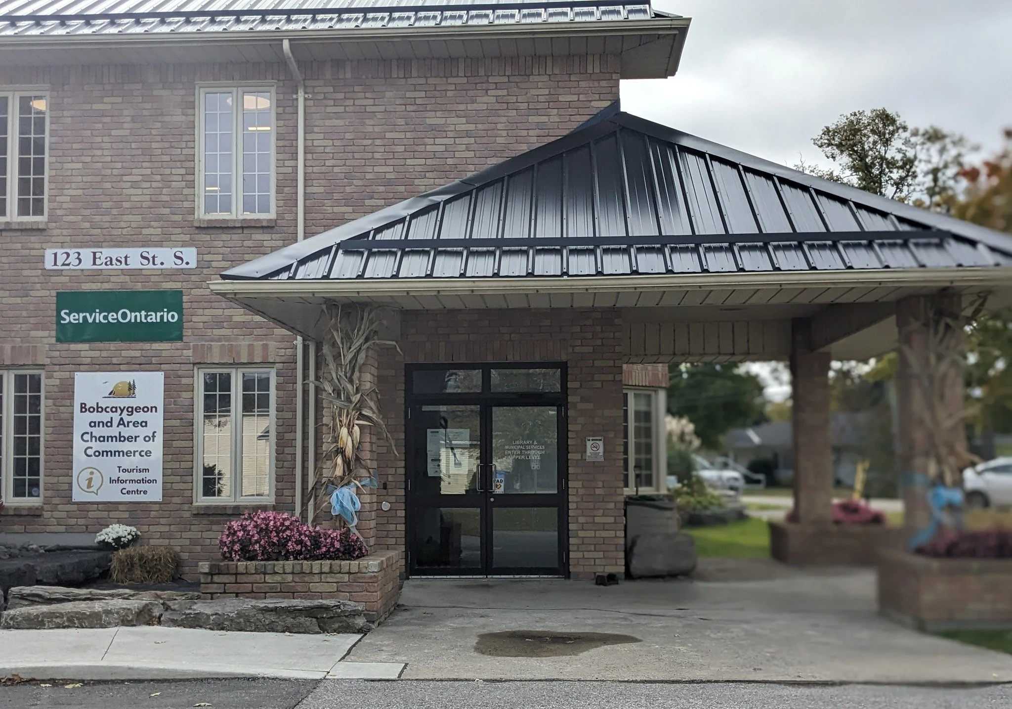 Front entrance of a brick building with signs indicating it is the Bobcaygeon and Area Chamber of Commerce, located at 123 East Street South in Ontario, with decorations including dried corn and flowers at the entrance.
