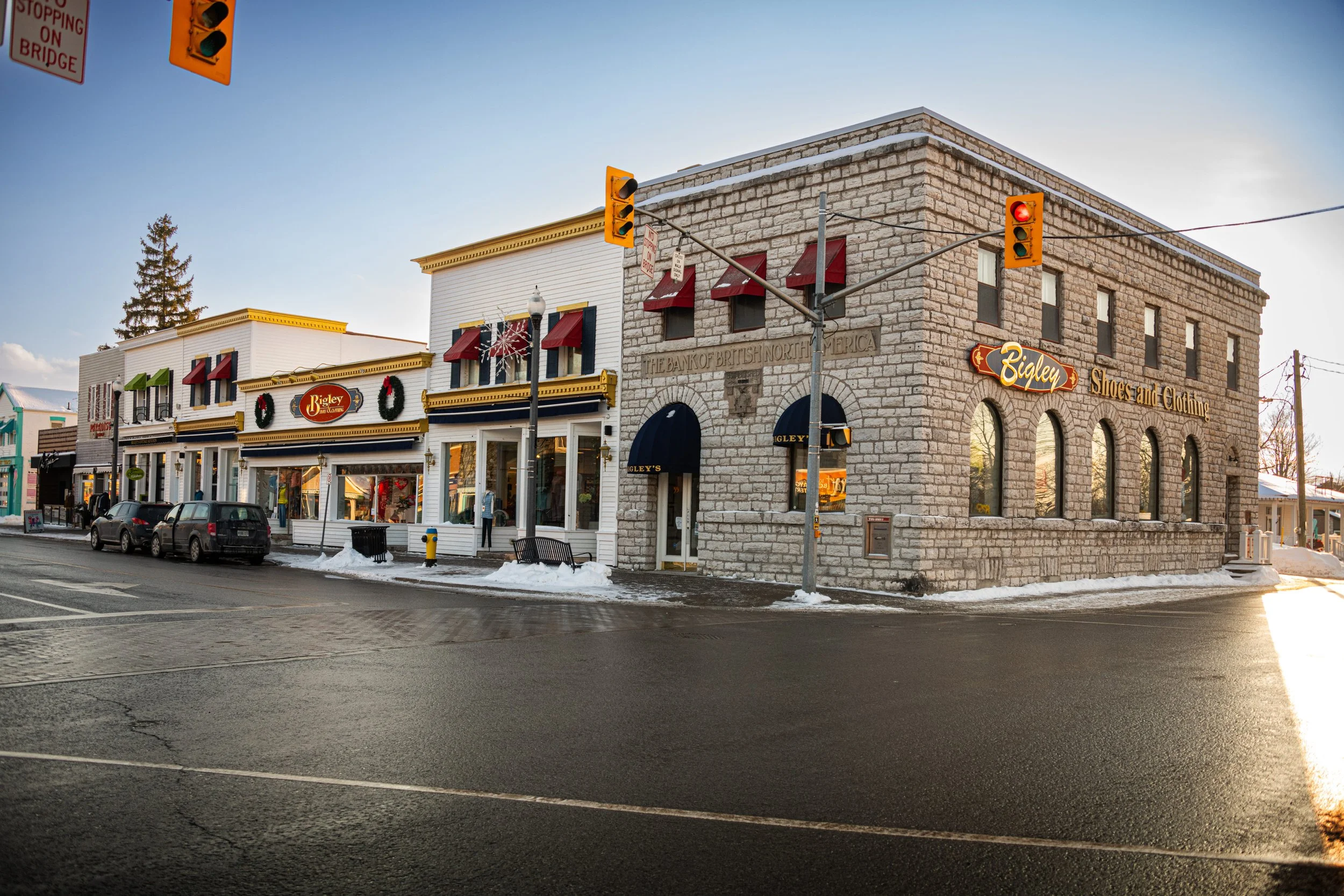 A street corner with a shopping district featuring buildings with signs that say "Bigley's Shoes and Clothing," decorated with holiday wreaths and red awnings, and streetlights. There are cars parked along the street with snow on the ground, and traffic signals at the intersection.