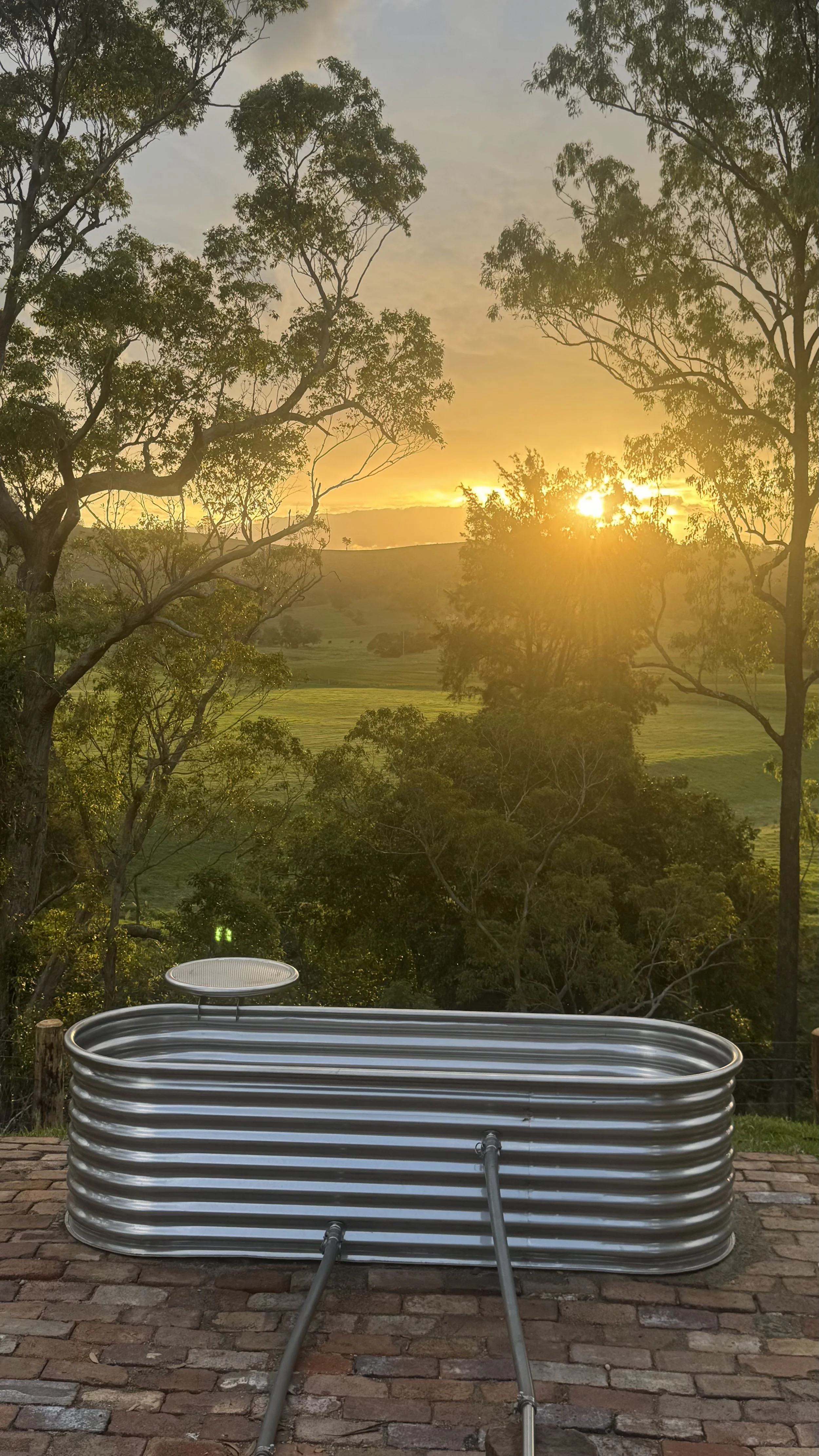 A large metal water tank on a brick patio with trees and a sunset in the background.