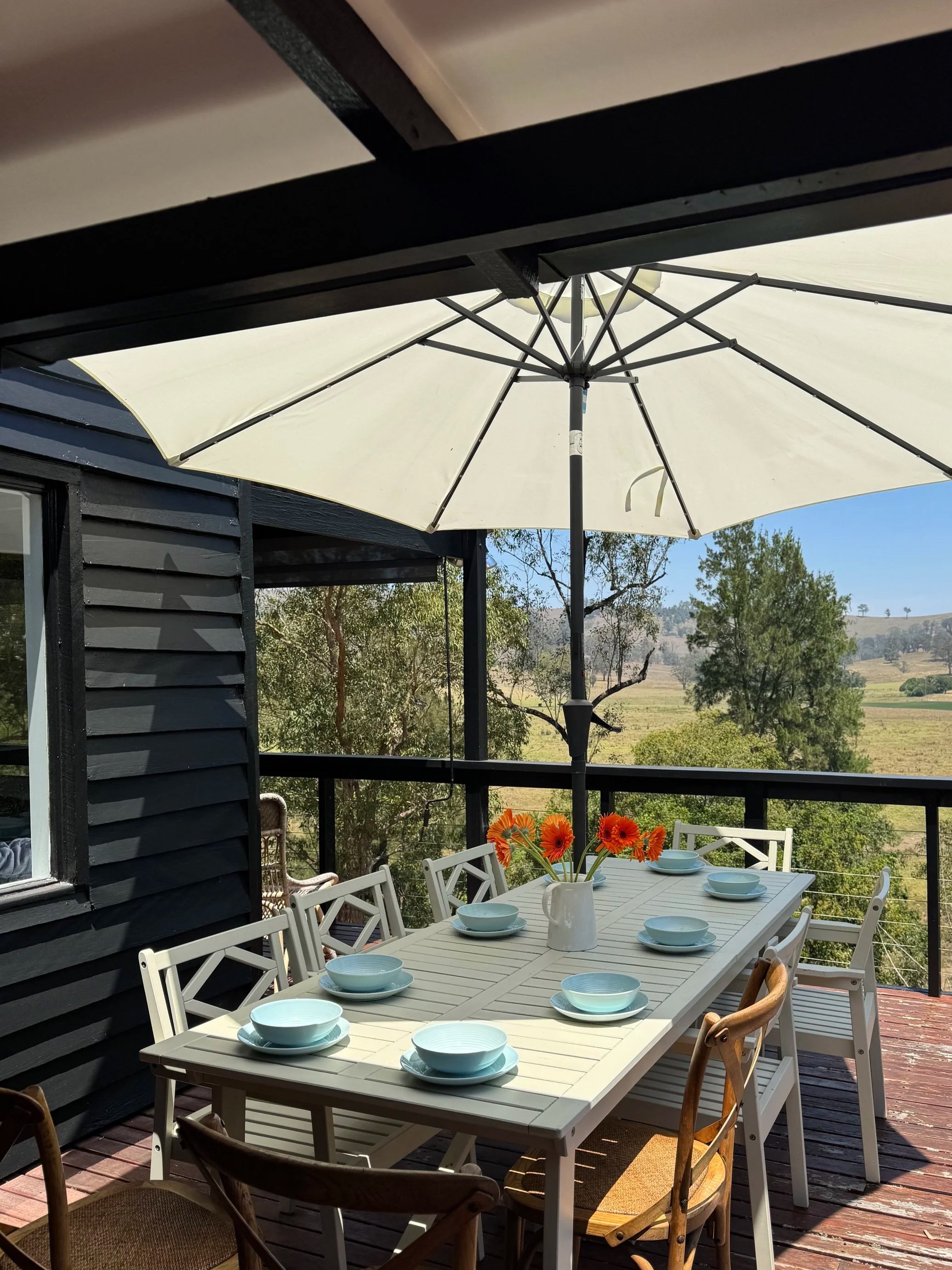 An outdoor patio with a white dining table set with 9 white bowls and a white pitcher with orange flowers, surrounded by mixed wood and white chairs, shaded by a large white umbrella, overlooking a scenic landscape with trees and fields.