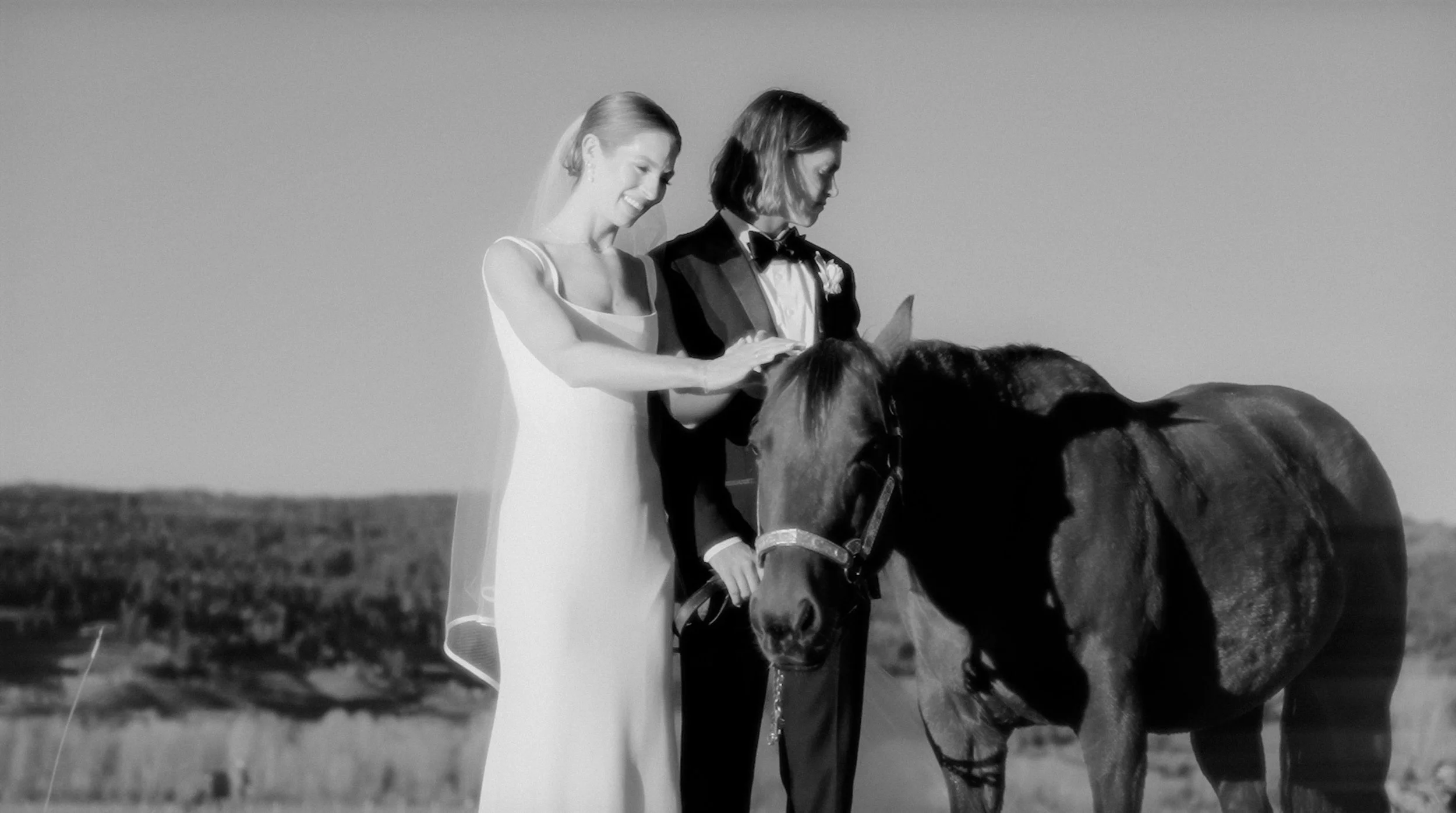 A black and white photo of a bride and groom standing outdoors with a horse. The bride is smiling and wearing a wedding dress, while the groom is dressed in a tuxedo and holding the horse's reins. The background shows a rural landscape.