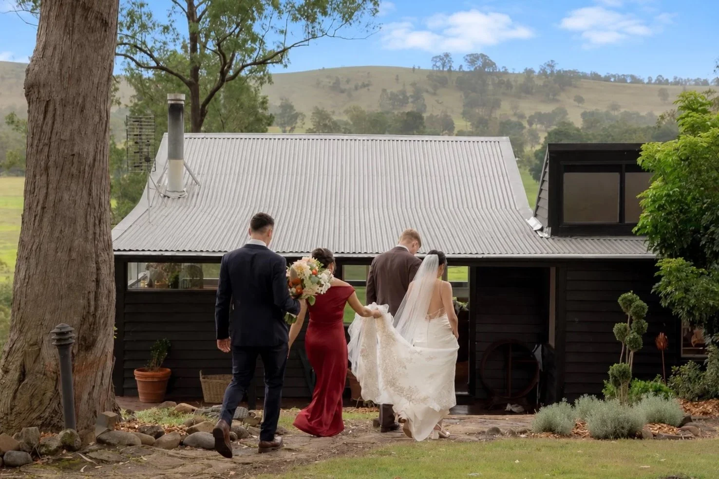 Bride, groom and bridesmaid walking toward the Kingaley farmhouse with rolling Hunter Valley hills in the background