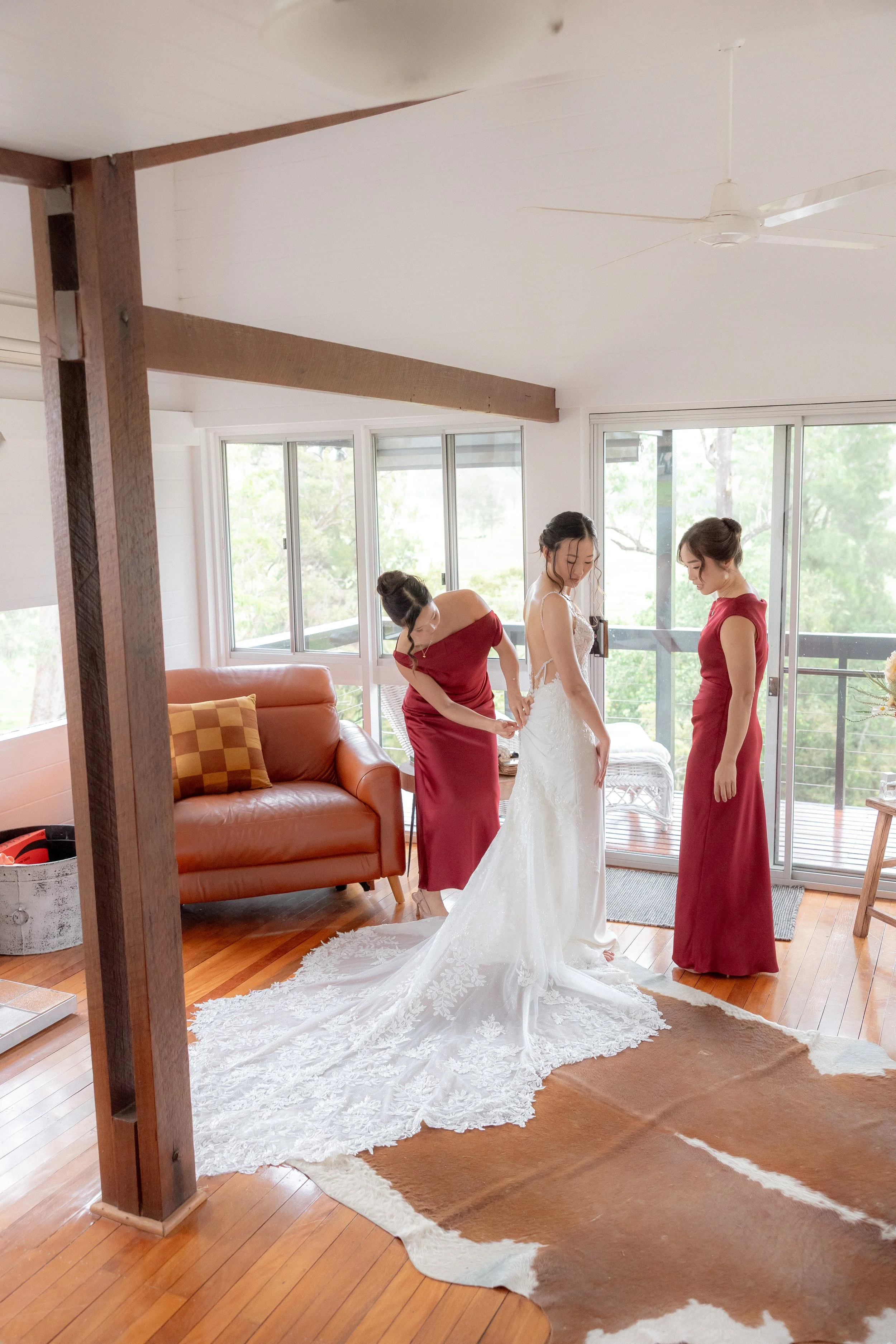 Bride having her wedding dress fastened by bridesmaids in red inside the Kingaley farmhouse before the ceremony, Dungog NSW