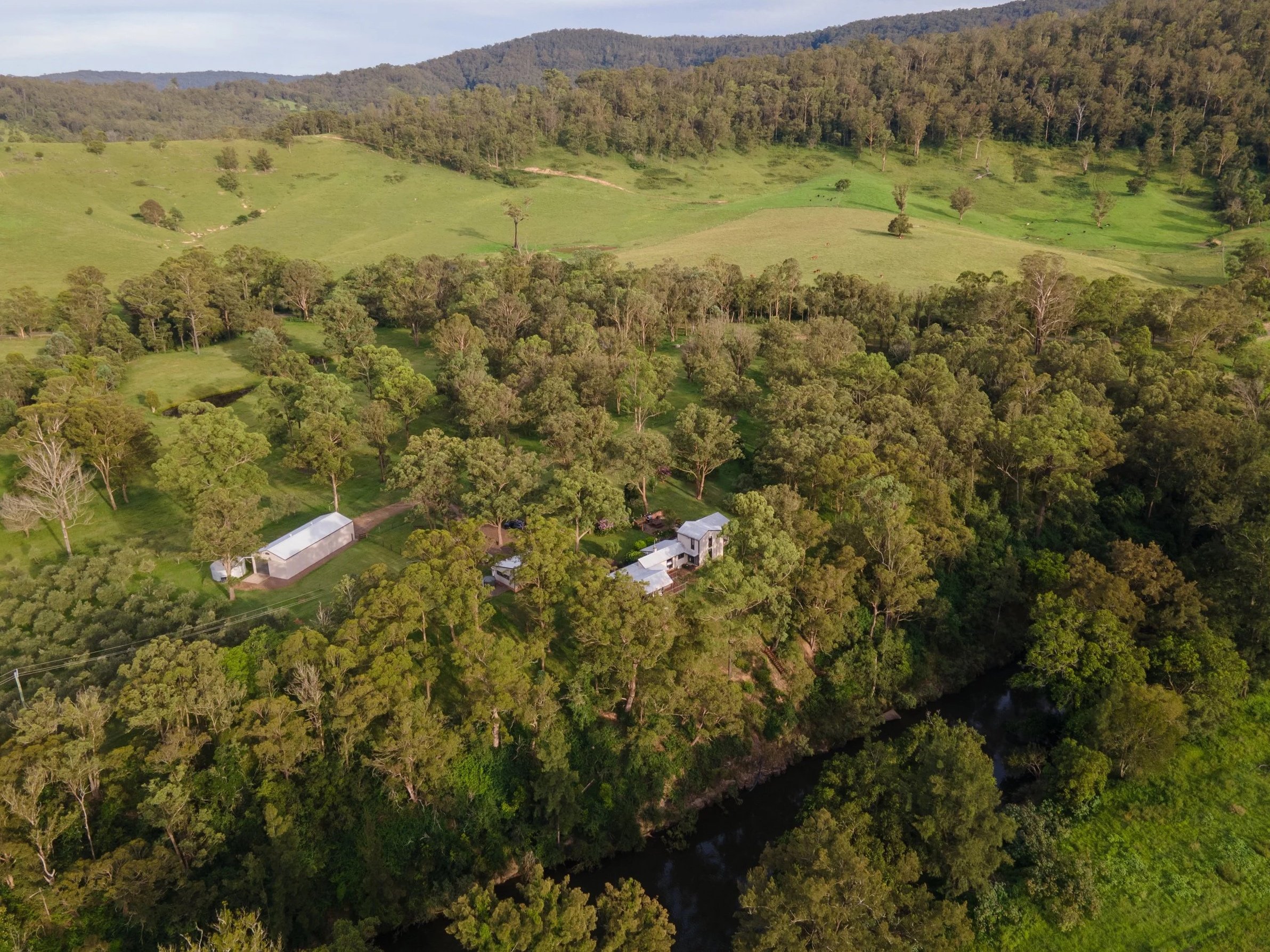 Aerial view of a lush, green rural landscape with rolling hills, trees, small buildings, and a winding river, under a partly cloudy sky.