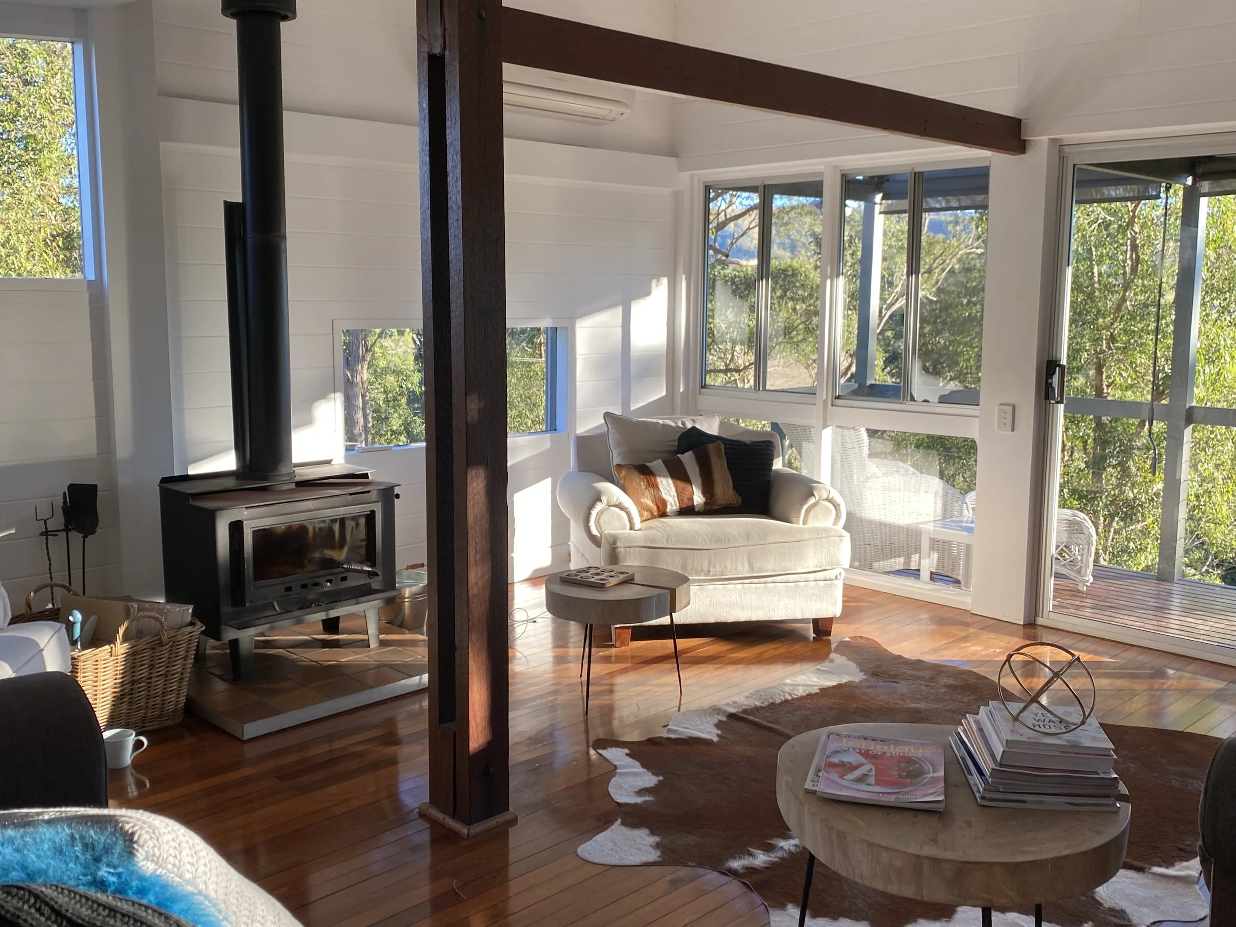 A bright living room with large windows and sliding glass door showing trees outside. There is a white armchair with pillows, a small round table with magazines, a wood stove, and a cowhide rug on a wooden floor.