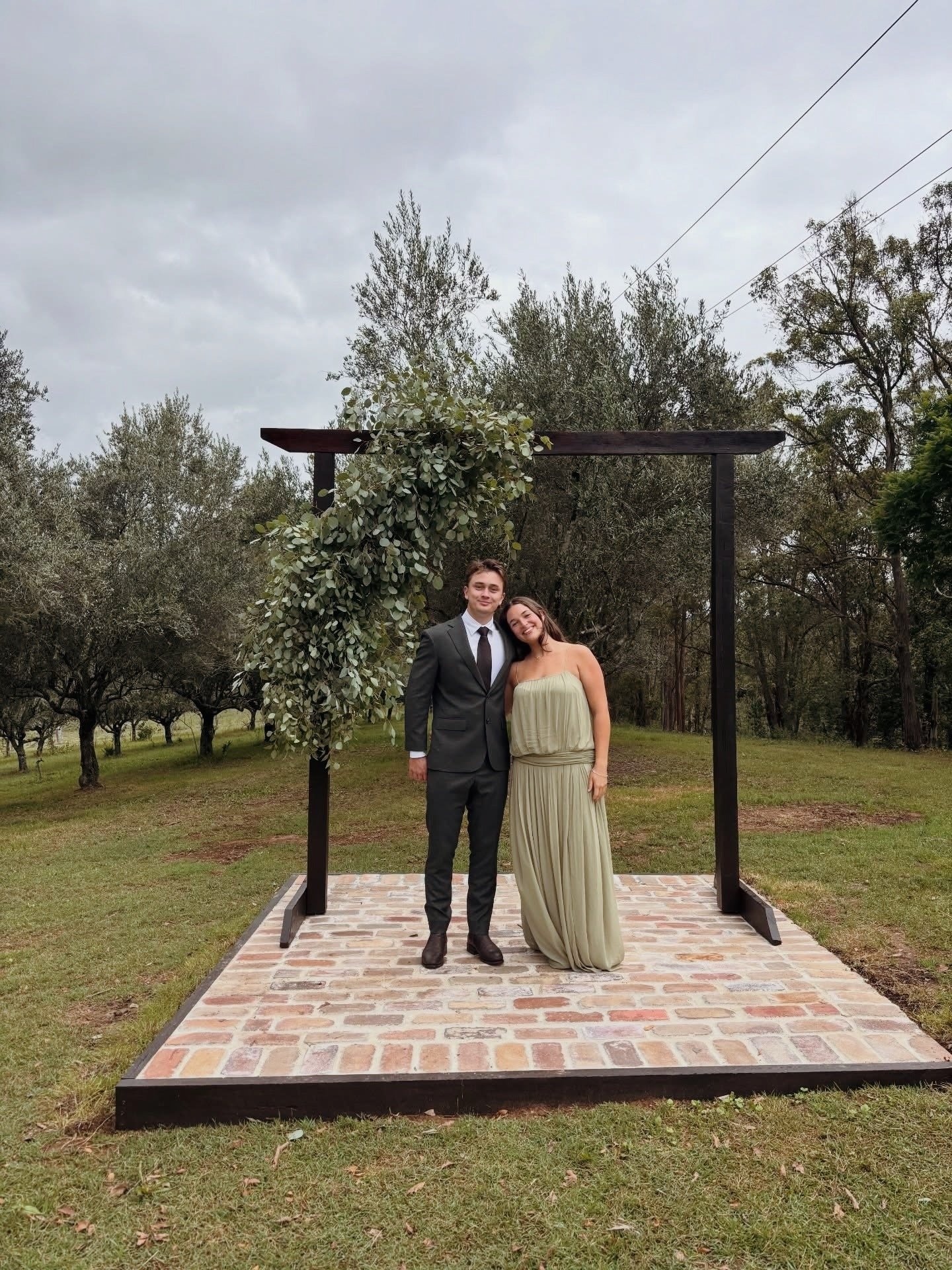A young couple dressed formally standing on a small brick platform outdoors under a black wooden arch decorated with green leafy foliage on a cloudy day.