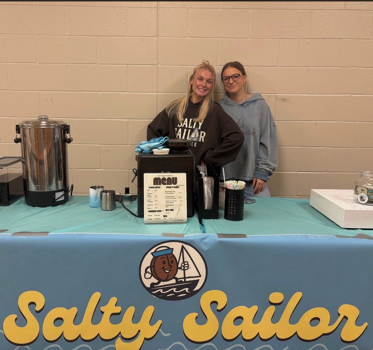 Two young women standing behind a beverage stand with a Salty Sailor banner in front. One woman is wearing a black Salty Sailor hoodie, and the other is wearing a gray Salty Sailor hoodie. The stand has a menu, a large coffee urn, cups, and straws.