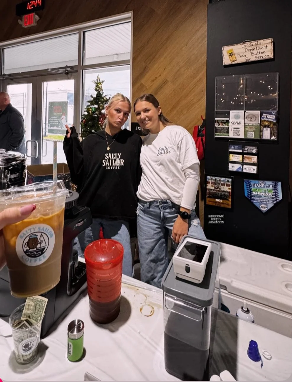 Two women posing inside a coffee shop, one wearing a black sweatshirt and the other a white sweatshirt, with a Christmas tree in the background. A hand holding a large iced coffee with a "Salty Sailor" logo is visible in the foreground. The shop has a wooden wall and a drink station on the counter.