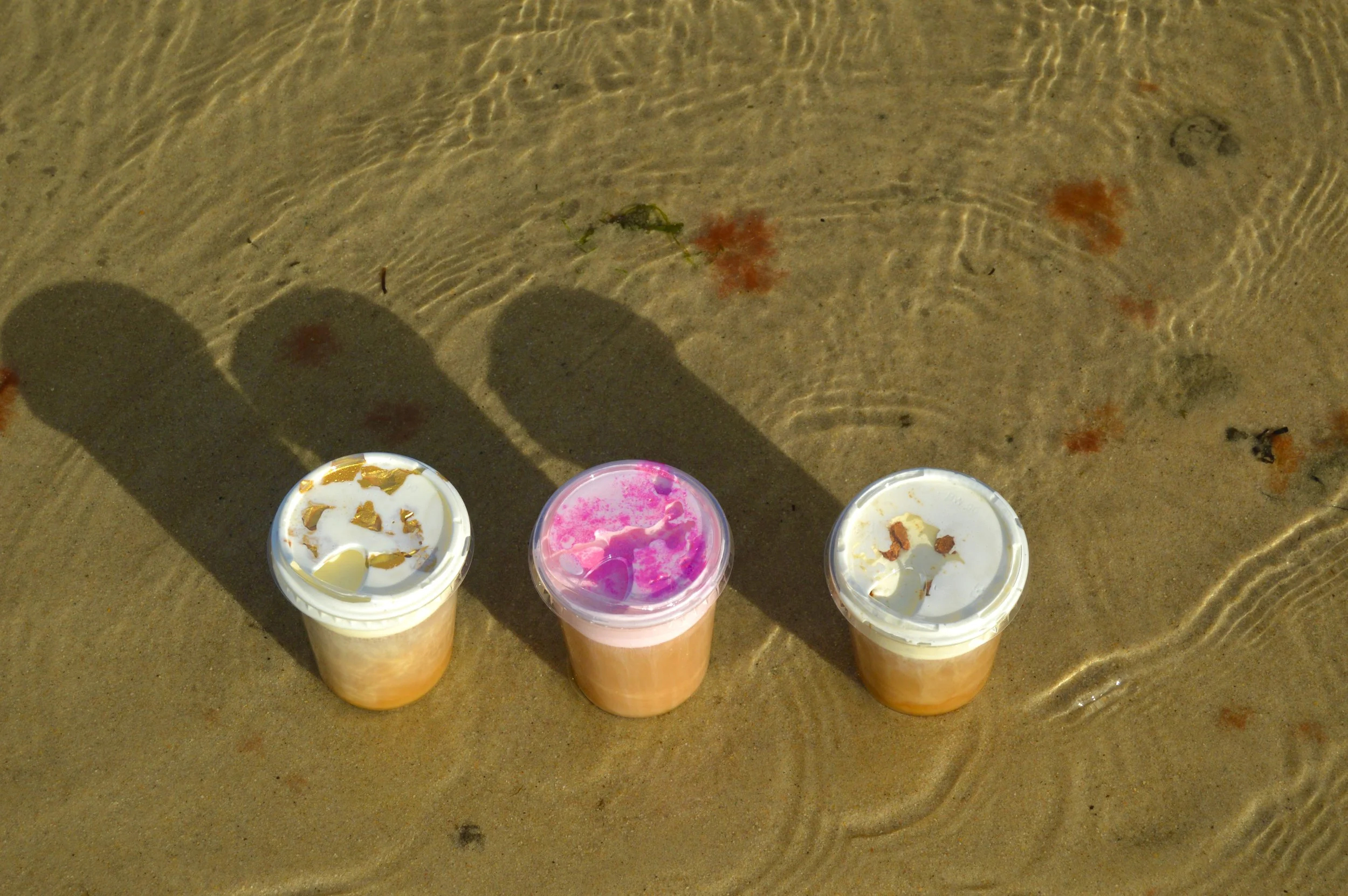 Three cups of drinks on the sandy beach, casting elongated shadows indicating bright sunlight, with some small plant debris and footprints in the sand