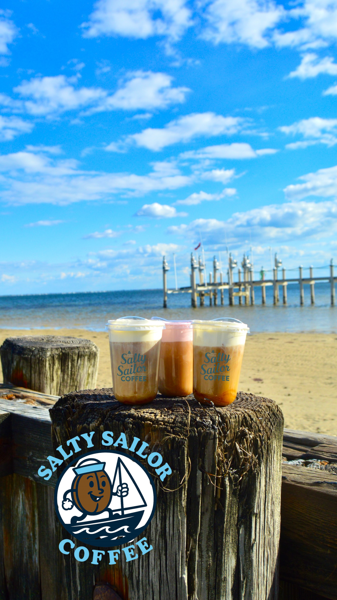 Three cups of coffee on a wooden post at the beach with a pier and blue sky with clouds in the background.