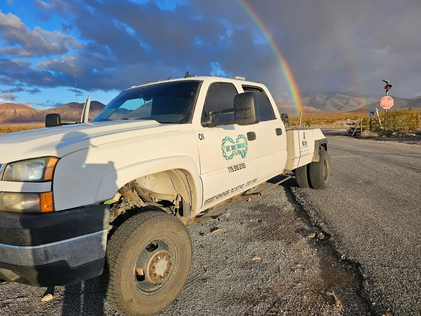A white utility truck parked on a gravel road in a desert landscape with mountains in the background. There is a vibrant rainbow in the sky and a stop sign on the right.