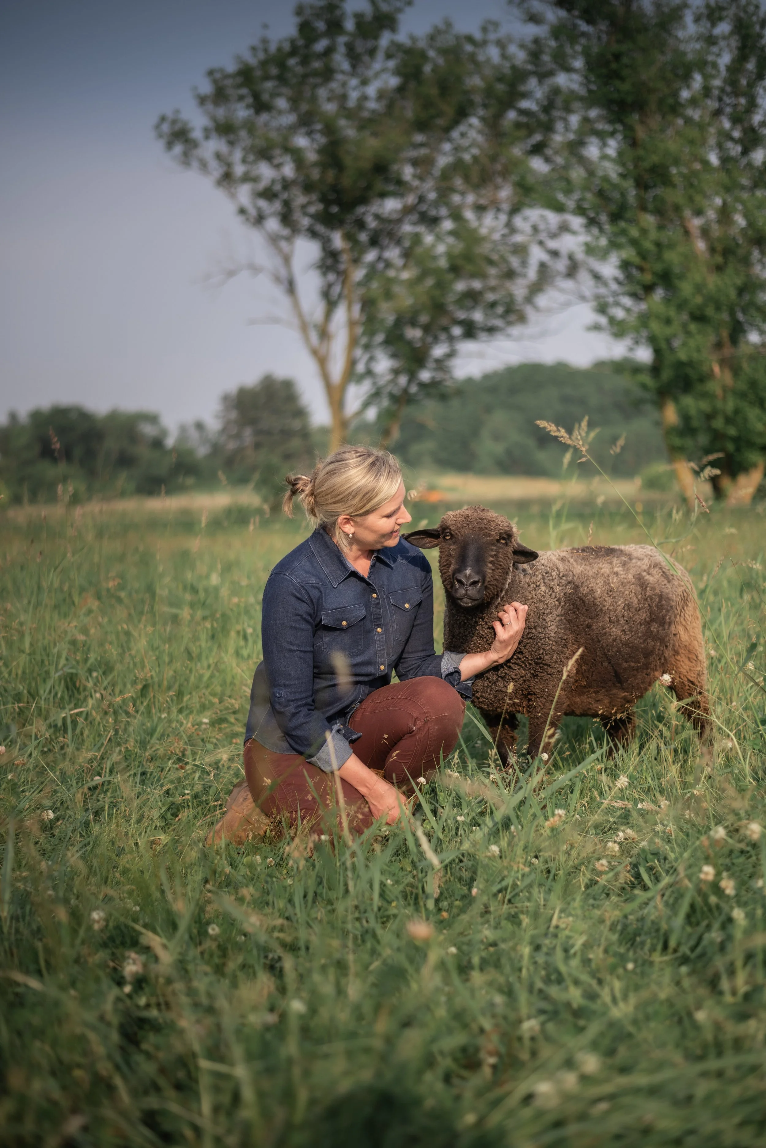 Woman kneeling in a grassy field petting a sheep, with trees in the background on a clear day.