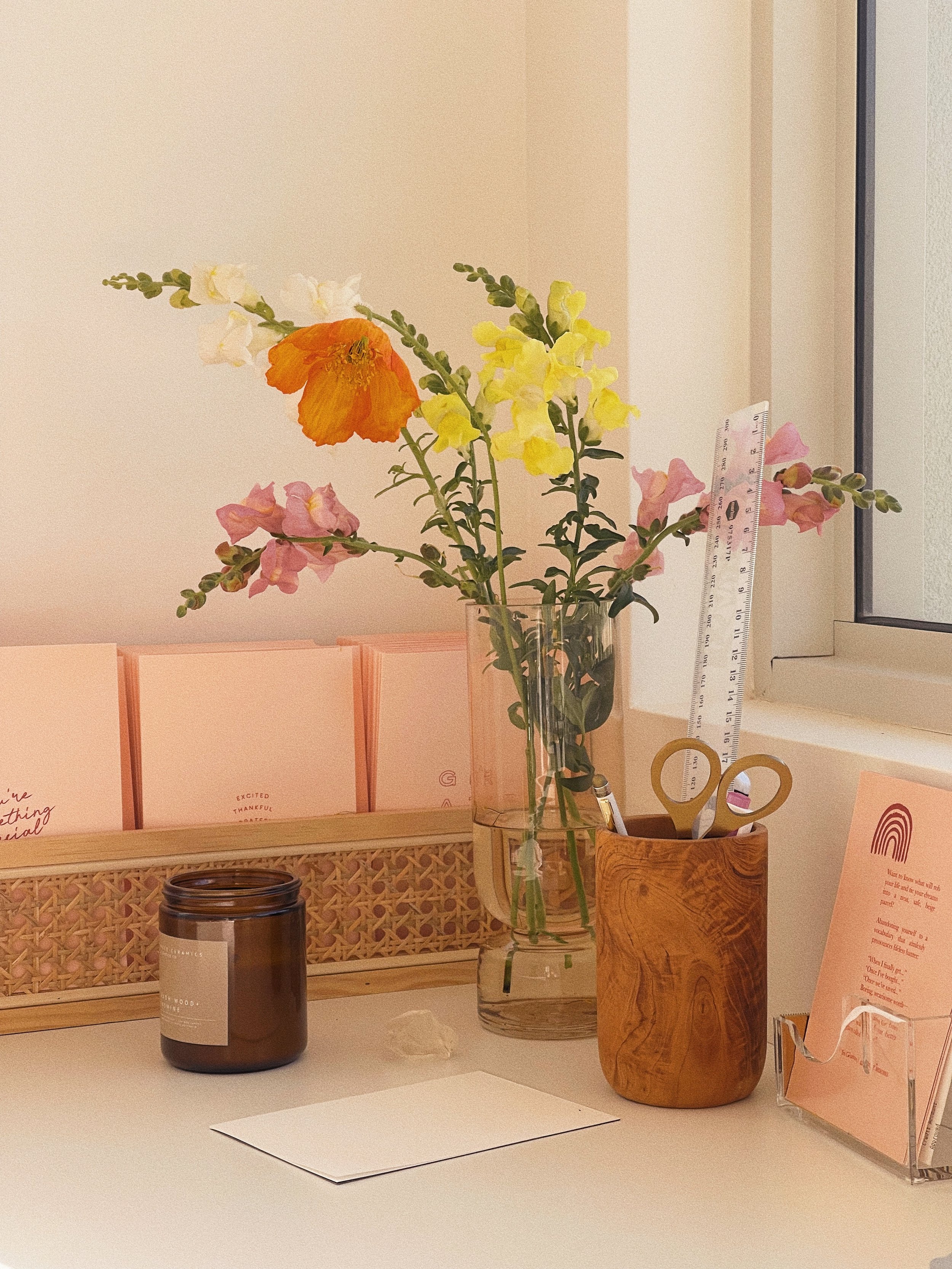 A desk with a glass vase of mixed flowers, a ruler, a wooden scissor holder with scissors, a candle, pink cards, and a pink info card holder near a window.
