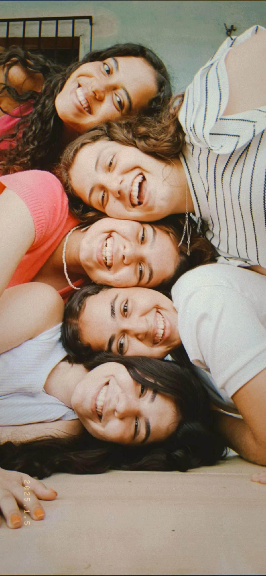 Six young women are lying on the floor, their heads close together, taking a selfie and smiling. They are indoors, with a metal door and some hooks in the background.