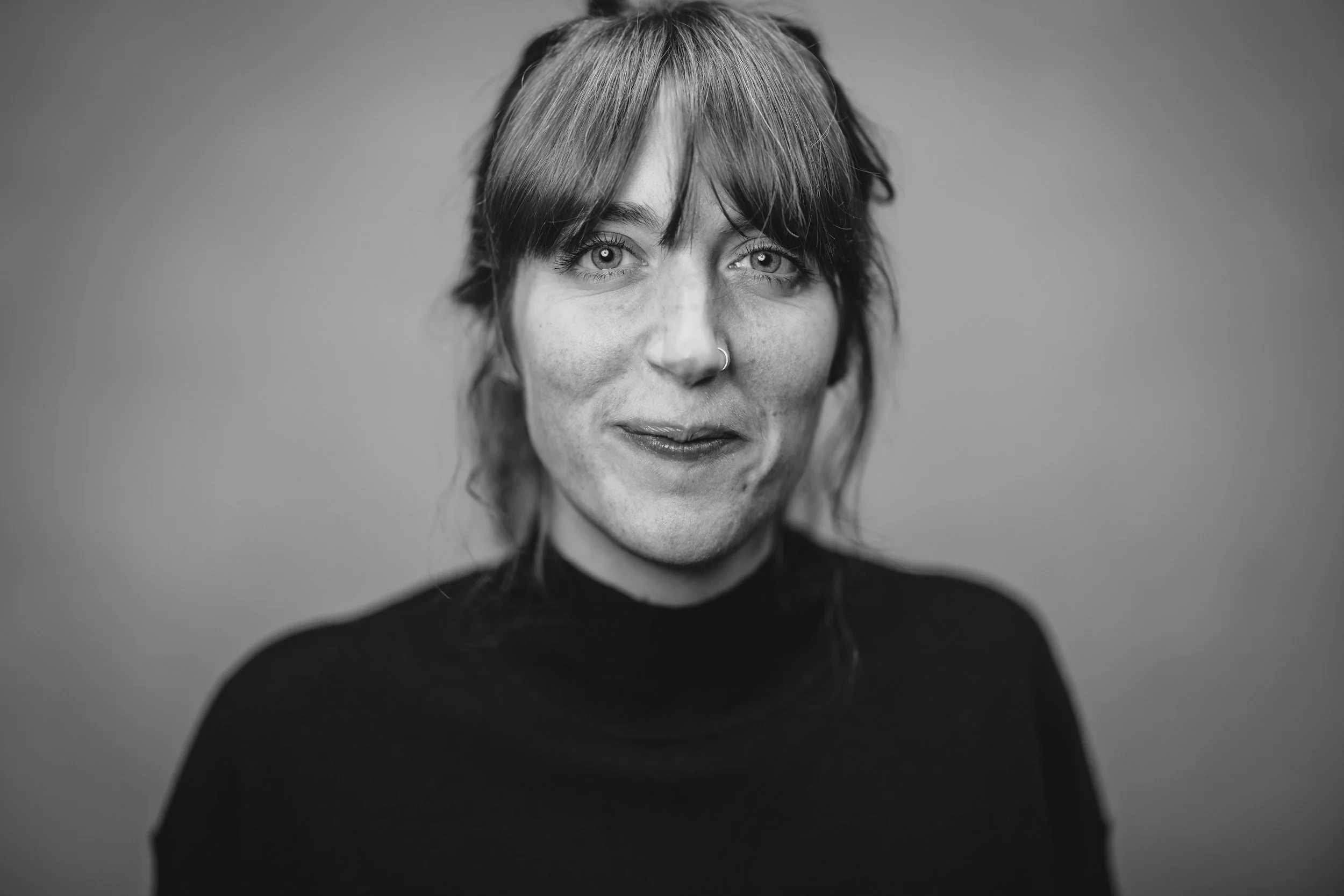 A woman with shoulder-length hair, wearing a nose ring, and a black turtleneck, smiling slightly at the camera, in black and white.