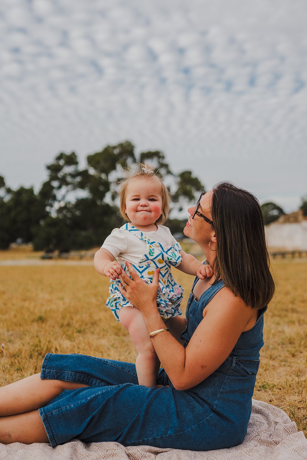 A woman sitting on a blanket outdoors, holding up a smiling toddler girl with blonde hair, wearing a white shirt and blue patterned shorts, against a background of grass, trees, and cloudy sky.